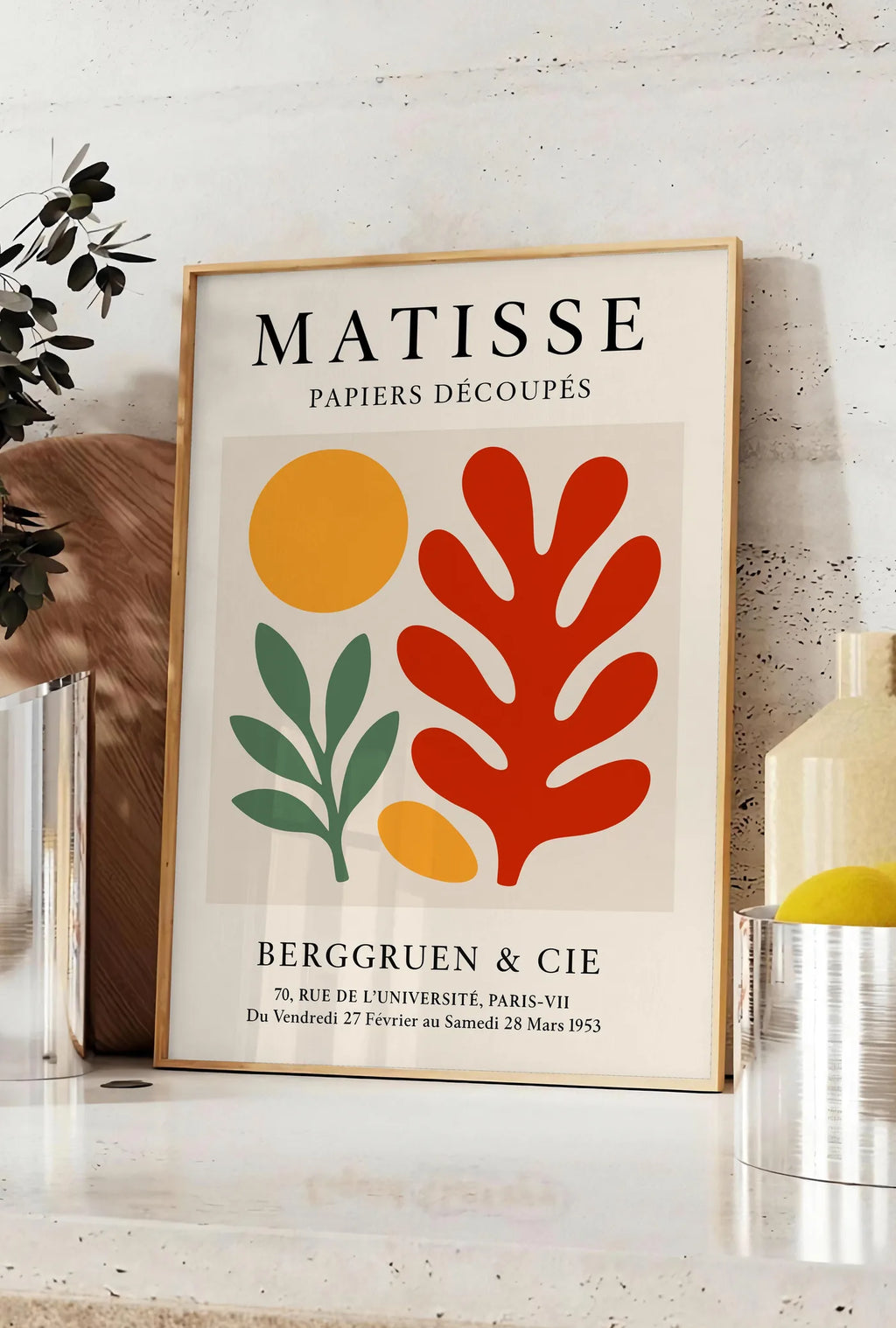 Affiche disque Branche et Corail Rouge en papiers decoupes Matisse sur fond colore, posee sur un plan de travail de cuisine en pierre claire, entre une branche d'eucalyptus en vase et un bocal de citrons