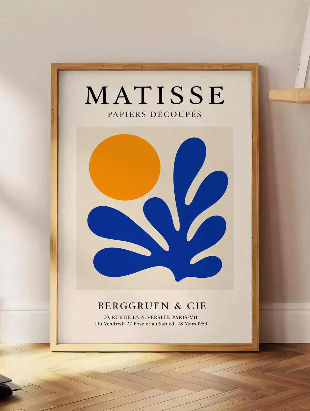 Estampe soleil Orange et Corail Bleu en papiers decoupes Matisse sur fond colore, posee au sol dans un coin salon, sur un parquet a chevrons, pres d'une table d'appoint en chene chargee de livres