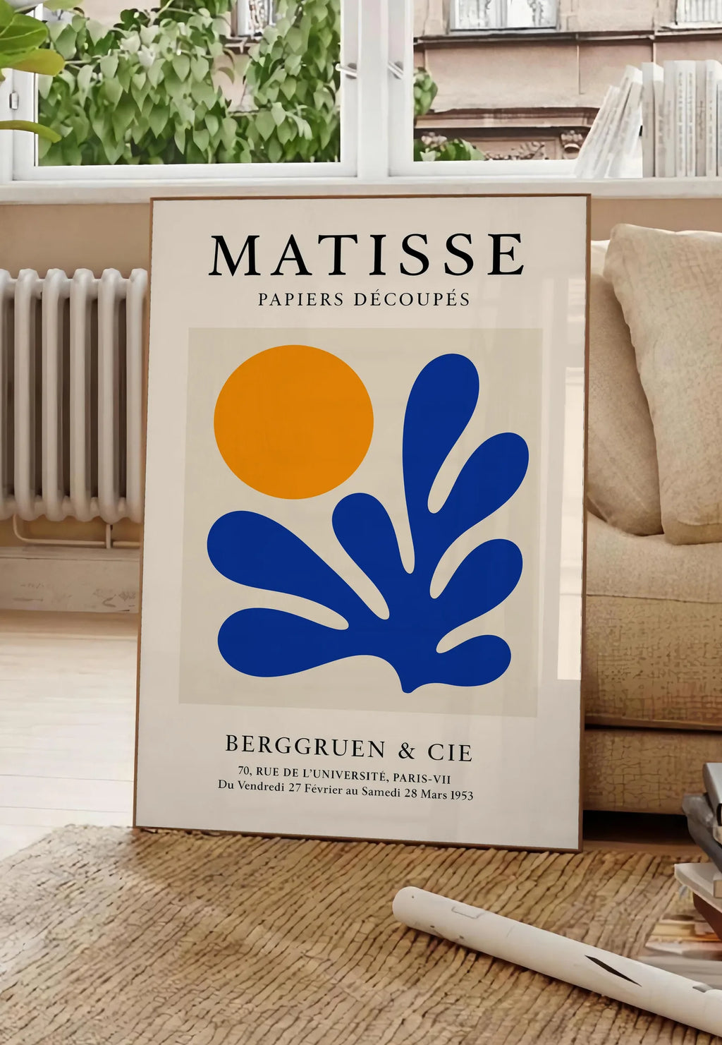 Poster soleil Orange et Corail Bleu en papiers decoupes Matisse sur fond colore, posee au sol dans un salon parisien, devant une grande fenetre donnant sur un immeuble haussmannien, entre un ficus et un canape creme