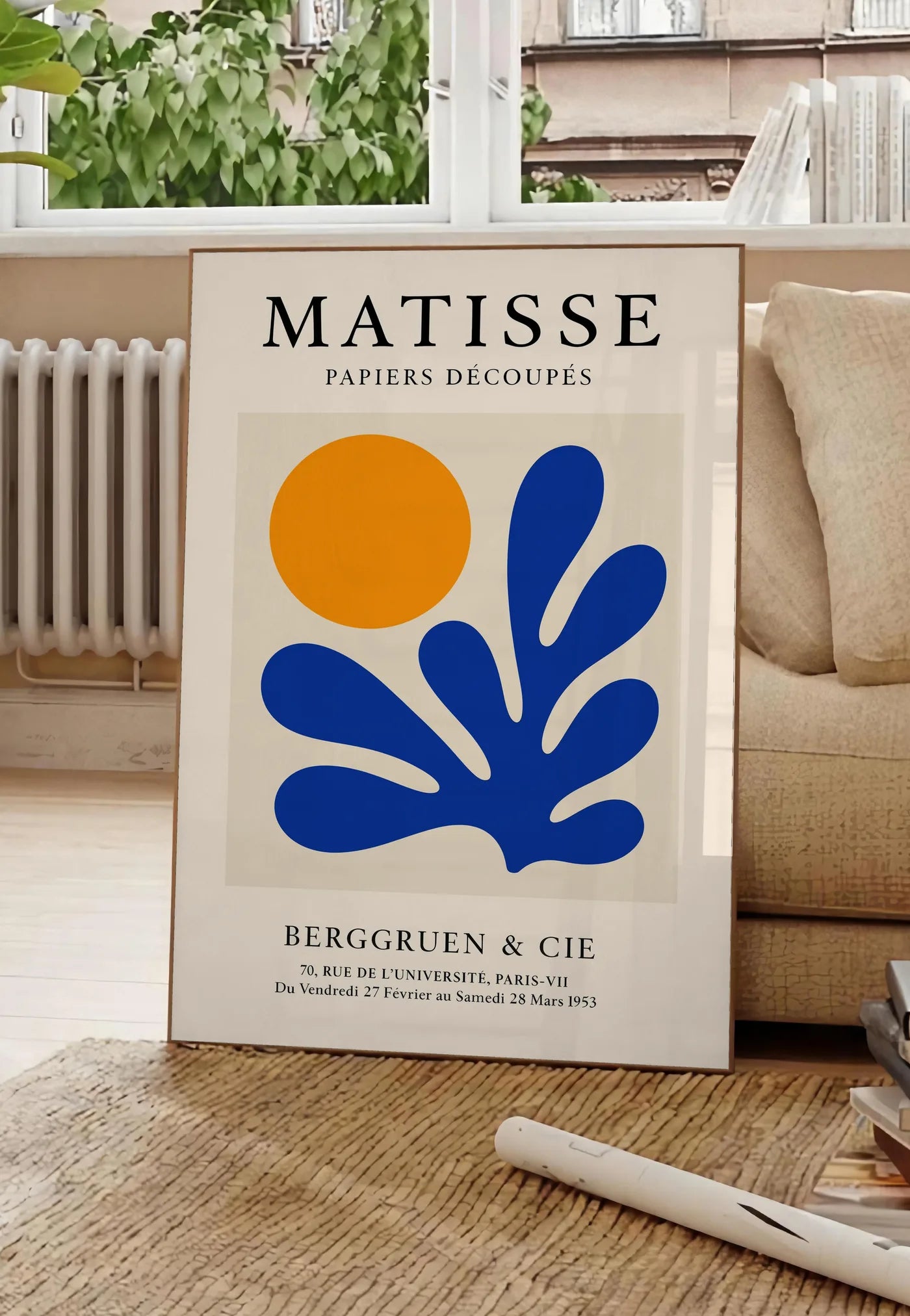 Poster soleil Orange et Corail Bleu en papiers decoupes Matisse sur fond colore, posee au sol dans un salon parisien, devant une grande fenetre donnant sur un immeuble haussmannien, entre un ficus et un canape creme