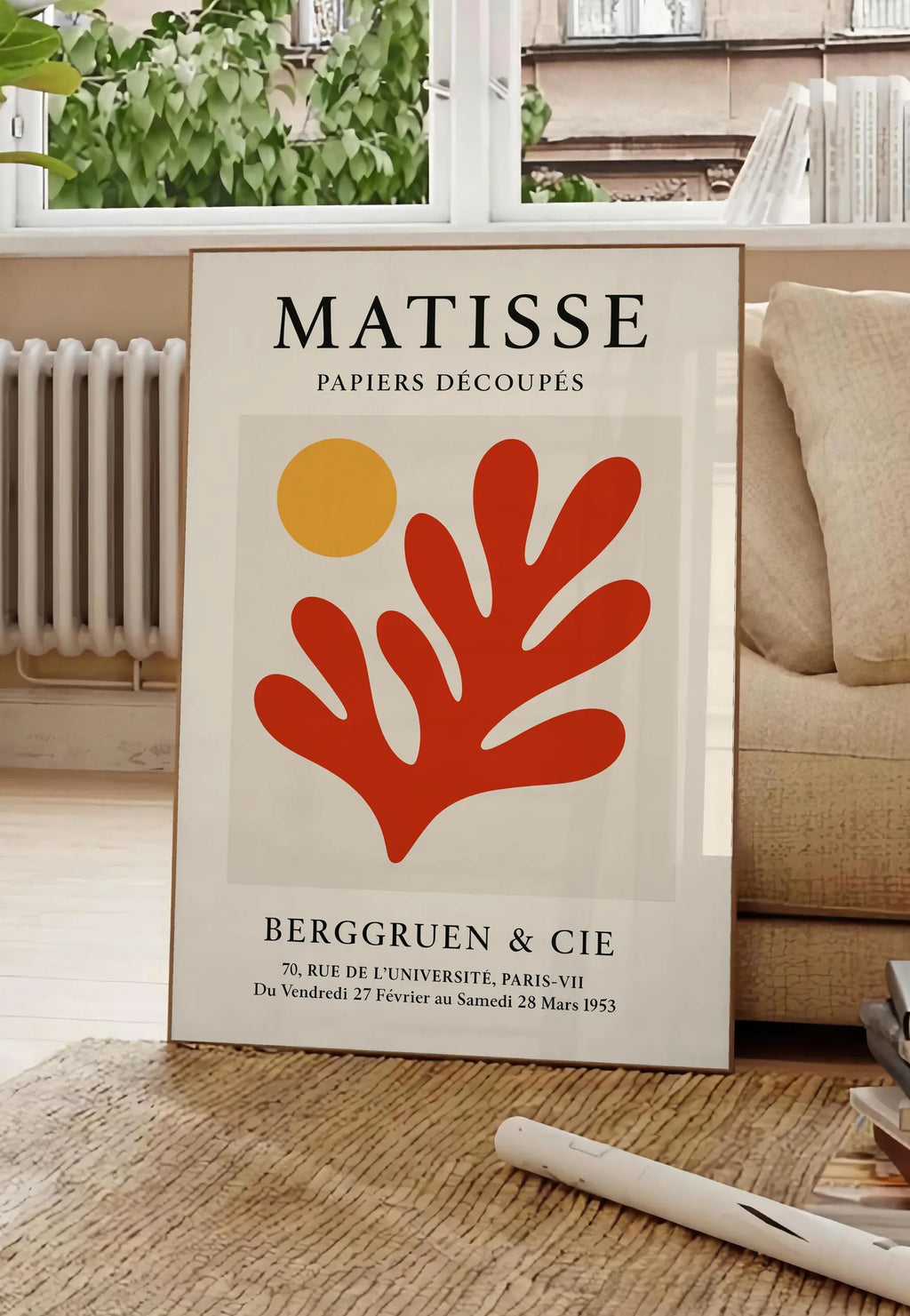 Poster soleil Orange et Corail Rouge en papiers decoupes Matisse sur fond colore, posee au sol dans un salon parisien, devant une grande fenetre donnant sur un immeuble haussmannien, entre un ficus et un canape creme