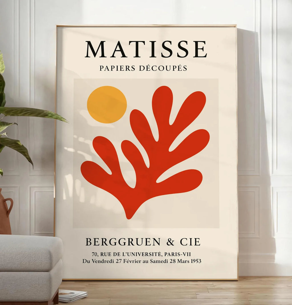 Reproduction soleil Orange et Corail Rouge en papiers decoupes Matisse sur fond colore, posee au sol dans un salon minimaliste clair, a cote d'un canape beige et d'une plante verte, sur un parquet clair