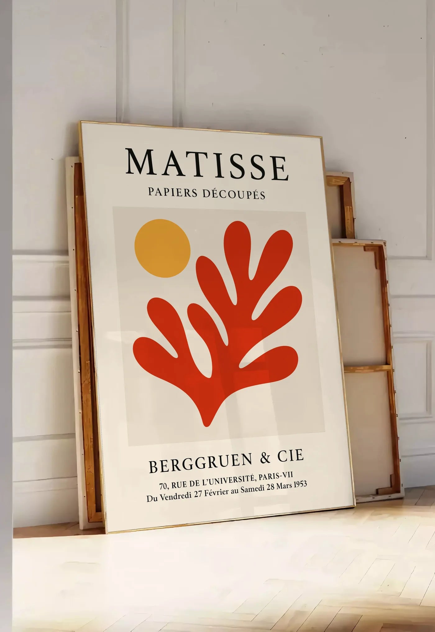 Tirage encadrable soleil Orange et Corail Rouge en papiers decoupes Matisse sur fond colore, posee au sol dans une entree lumineuse, contre un mur a panneaux blancs, a cote de chassis en bois empiles