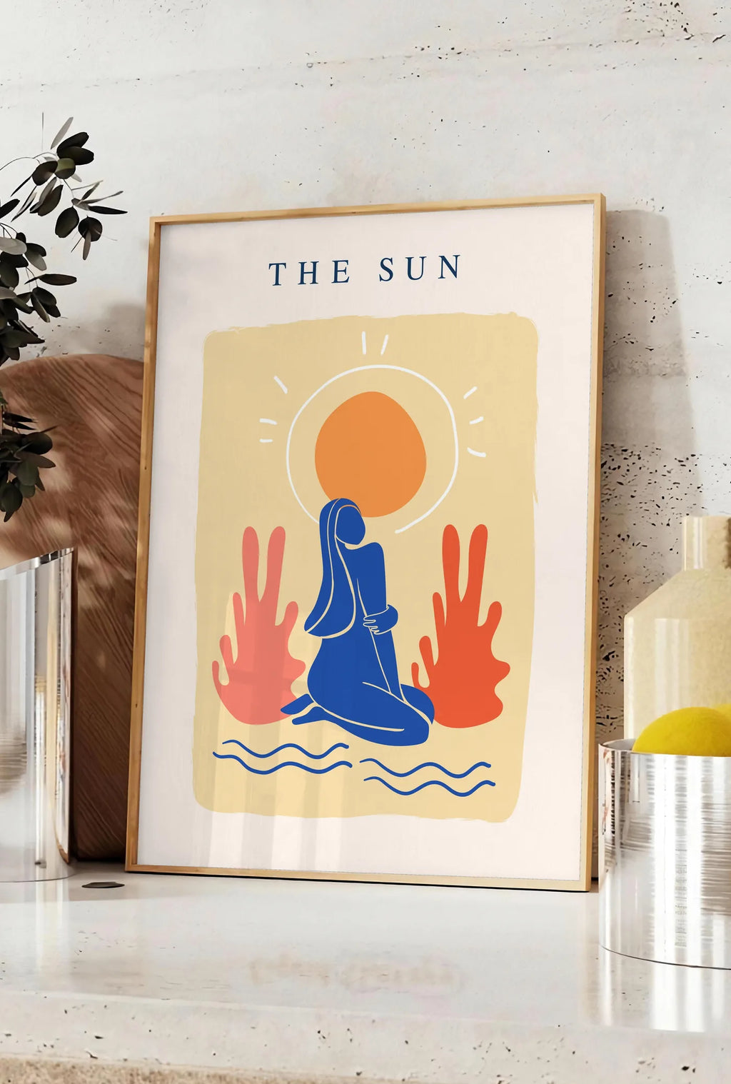 Affiche femme bleue agenouillee devant un soleil orange et des plantes corail, posee sur un plan de travail de cuisine en pierre claire, entre une branche d'eucalyptus en vase et un bocal de citrons