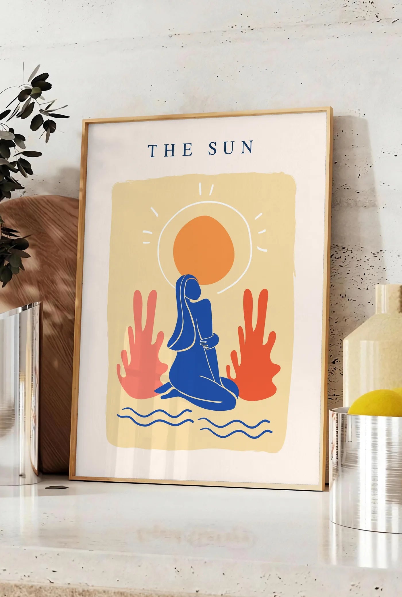 Affiche femme bleue agenouillee devant un soleil orange et des plantes corail, posee sur un plan de travail de cuisine en pierre claire, entre une branche d'eucalyptus en vase et un bocal de citrons