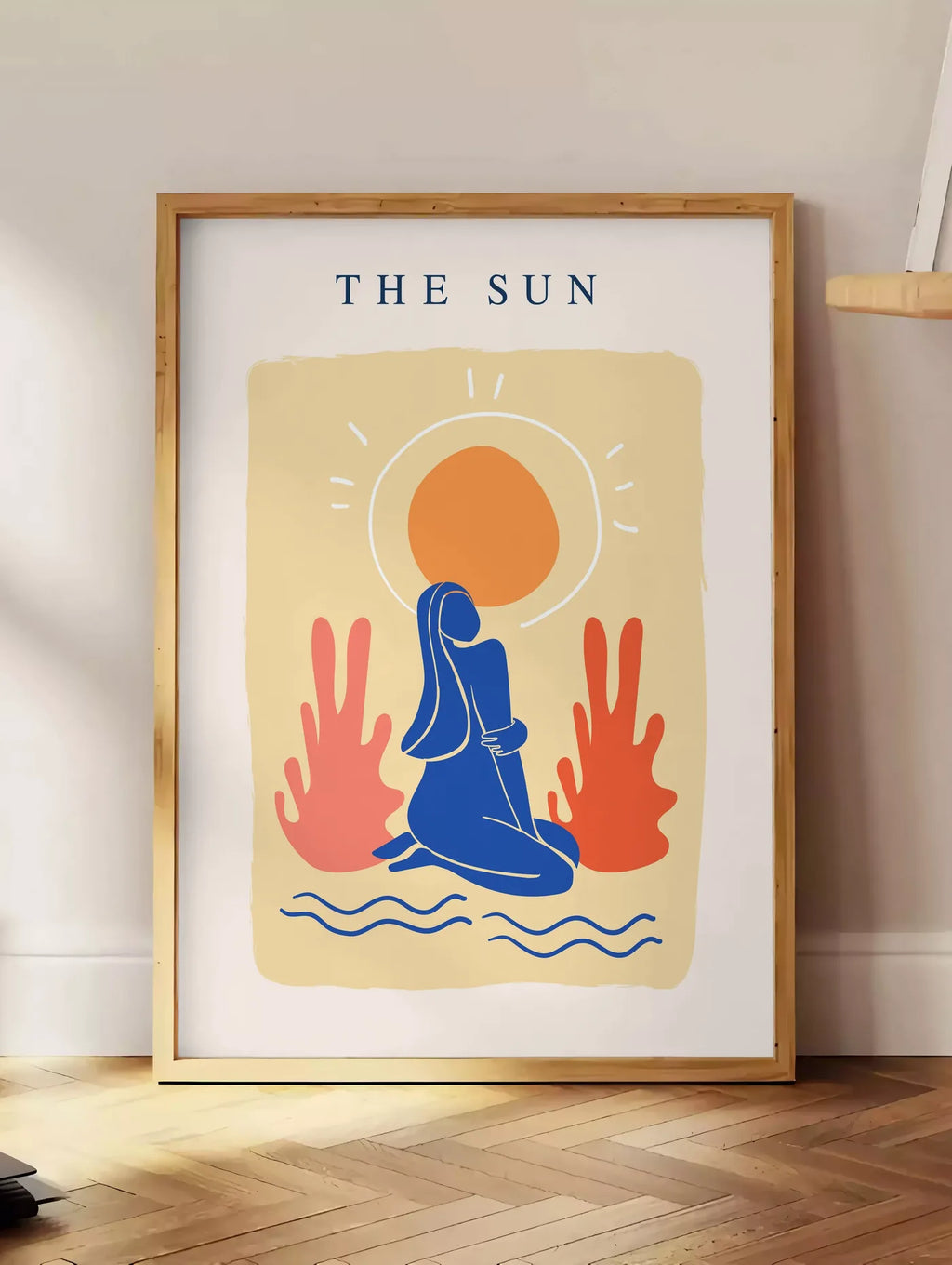 Estampe femme bleue agenouillee devant un soleil orange et des plantes corail, posee au sol dans un coin salon, sur un parquet a chevrons, pres d'une table d'appoint en chene chargee de livres