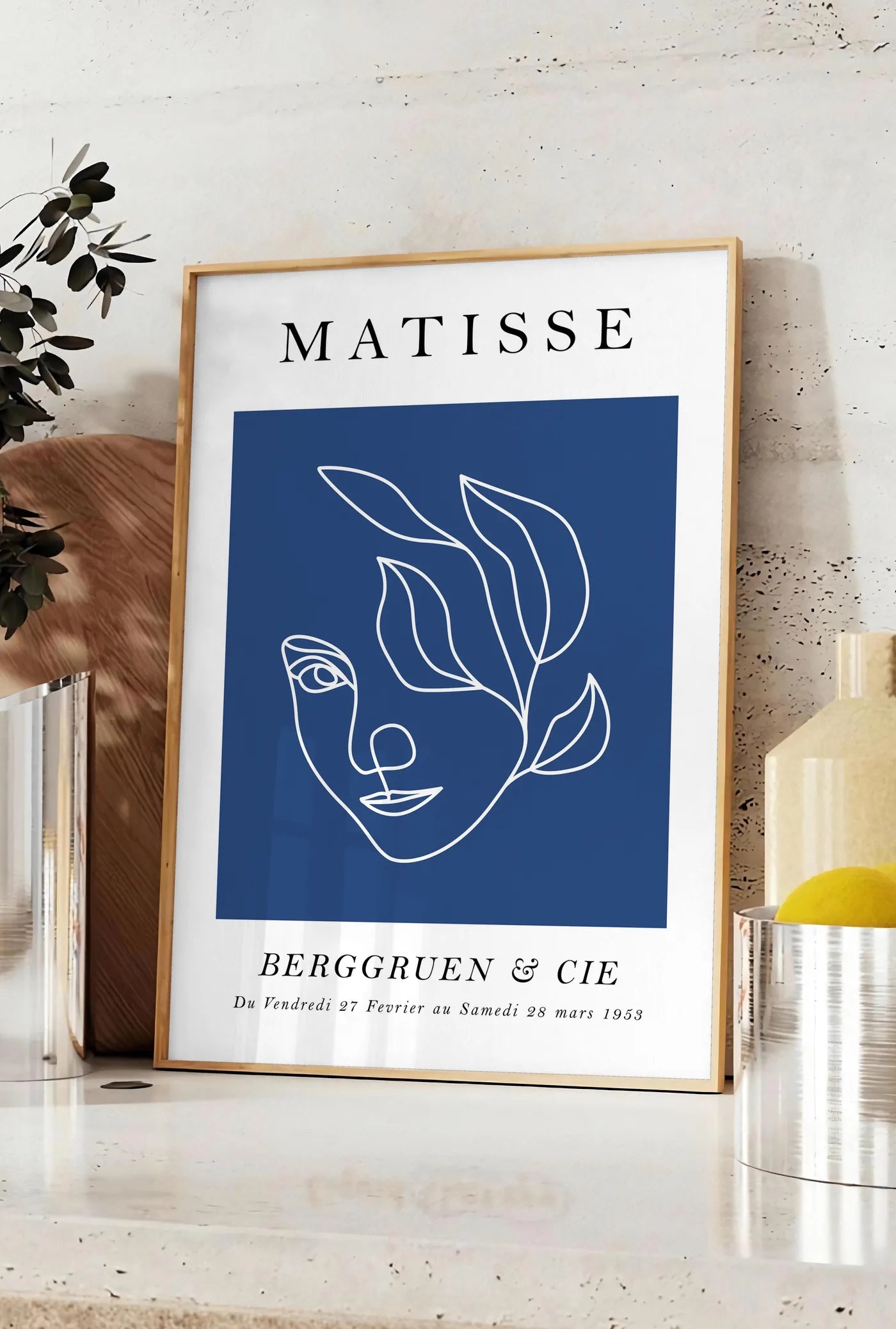 Affiche visage feminin au trait continu blanc sur fond bleu, posee sur un plan de travail de cuisine en pierre claire, entre une branche d'eucalyptus en vase et un bocal de citrons