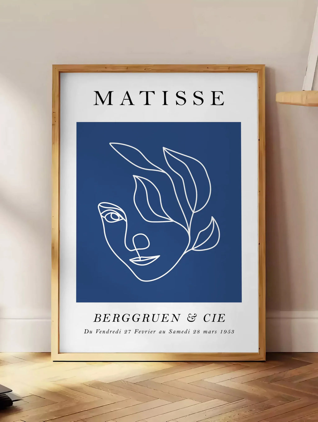 Estampe visage feminin au trait continu blanc sur fond bleu, posee au sol dans un coin salon, sur un parquet a chevrons, pres d'une table d'appoint en chene chargee de livres
