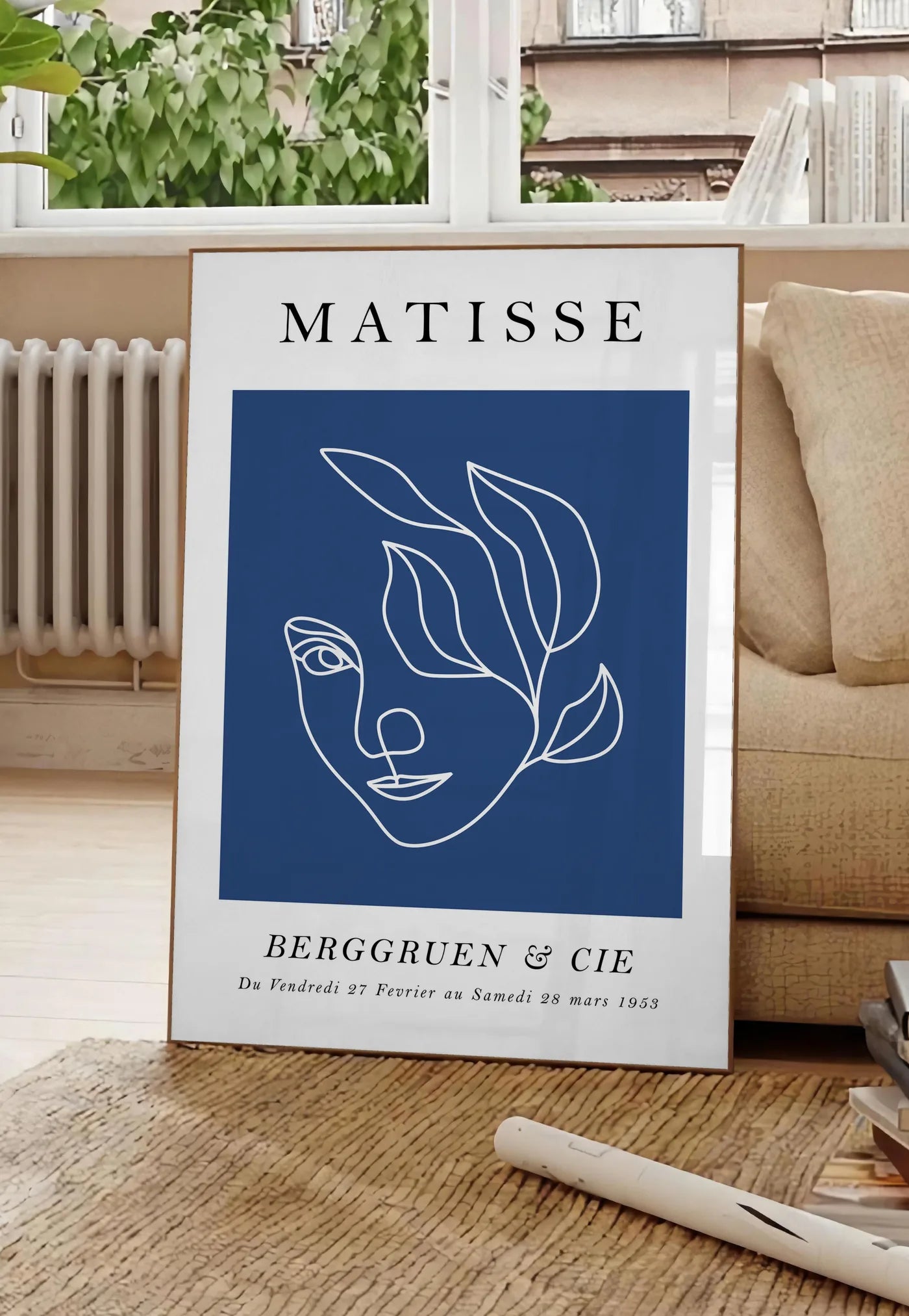 Poster visage feminin au trait continu blanc sur fond bleu, posee au sol dans un salon parisien, devant une grande fenetre donnant sur un immeuble haussmannien, entre un ficus et un canape creme