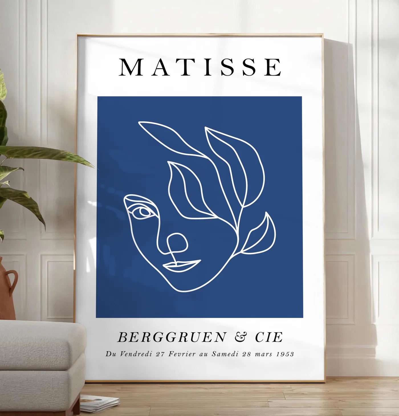 Reproduction visage feminin au trait continu blanc sur fond bleu, posee au sol dans un salon minimaliste clair, a cote d'un canape beige et d'une plante verte, sur un parquet clair