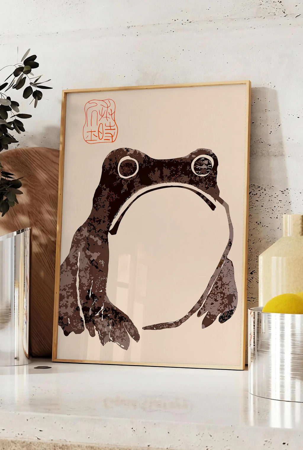 Affiche affiche grenouille blasee de Matsumoto Hoji, posee sur un plan de travail de cuisine en pierre claire, entre une branche d'eucalyptus en vase et un bocal de citrons