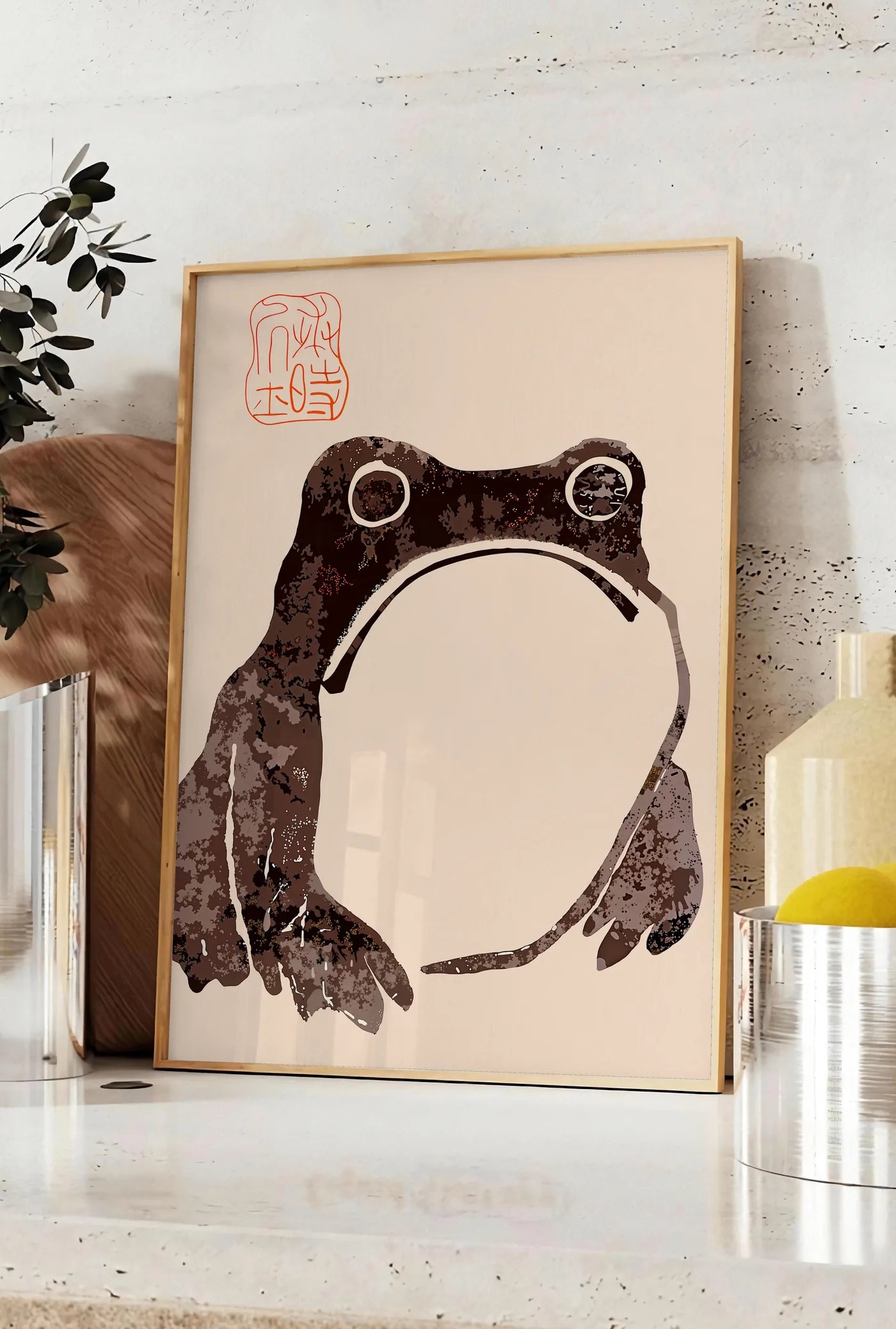 Affiche affiche grenouille blasee de Matsumoto Hoji, posee sur un plan de travail de cuisine en pierre claire, entre une branche d'eucalyptus en vase et un bocal de citrons