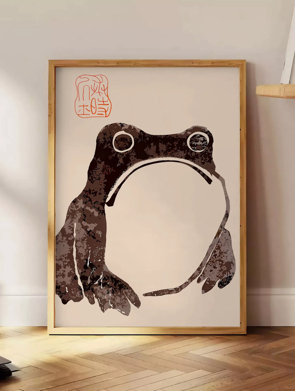 Estampe affiche grenouille blasee de Matsumoto Hoji, posee au sol dans un coin salon, sur un parquet a chevrons, pres d'une table d'appoint en chene chargee de livres