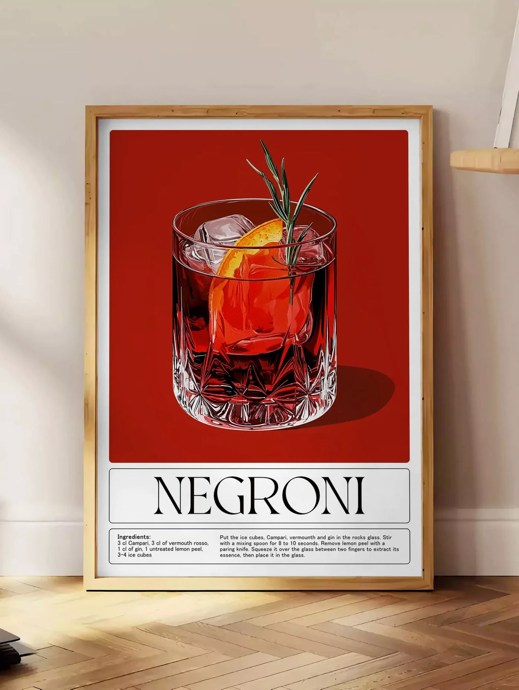 Estampe affiche cocktail Negroni, posee au sol dans un coin salon, sur un parquet a chevrons, pres d'une table d'appoint en chene chargee de livres