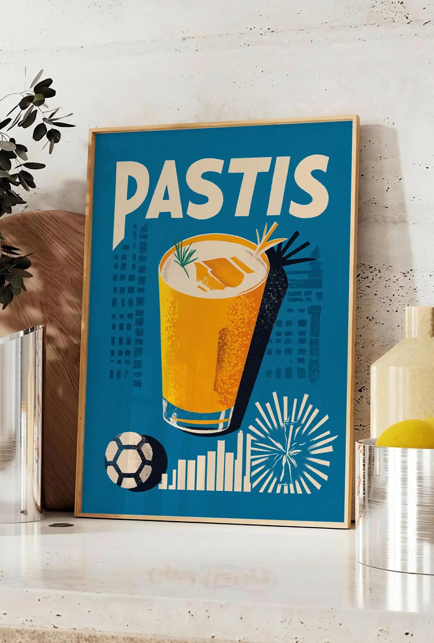Affiche affiche cocktail Pastis Marseille, posee sur un plan de travail de cuisine en pierre claire, entre une branche d'eucalyptus en vase et un bocal de citrons