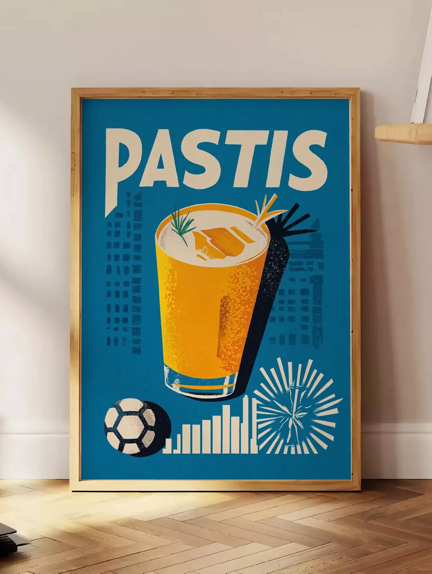 Estampe affiche cocktail Pastis Marseille, posee au sol dans un coin salon, sur un parquet a chevrons, pres d'une table d'appoint en chene chargee de livres