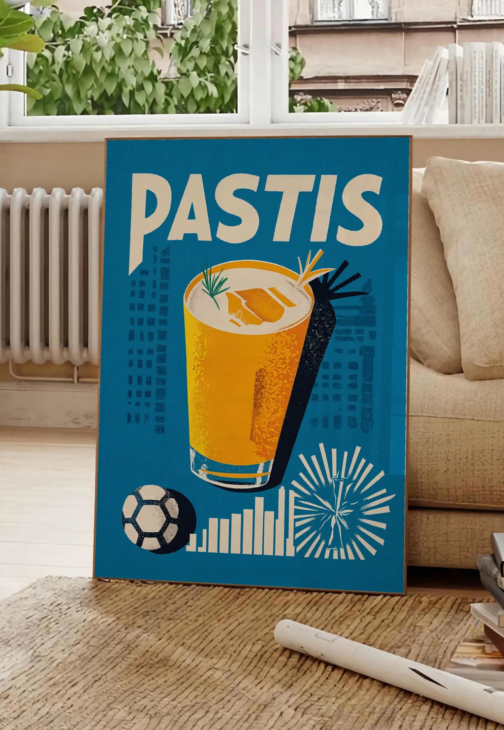 Poster affiche cocktail Pastis Marseille, posee au sol dans un salon parisien, devant une grande fenetre donnant sur un immeuble haussmannien, entre un ficus et un canape creme
