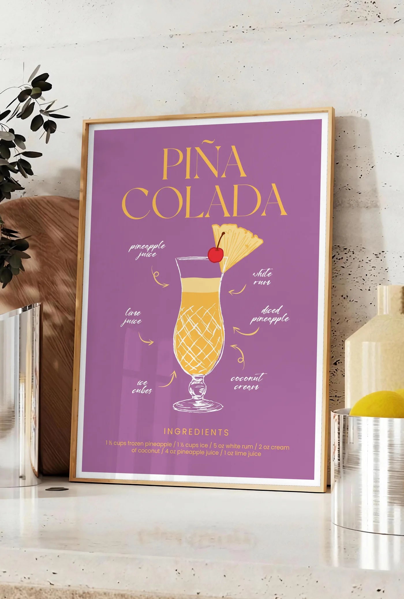 Affiche affiche cocktail Pina Colada, posee sur un plan de travail de cuisine en pierre claire, entre une branche d'eucalyptus en vase et un bocal de citrons