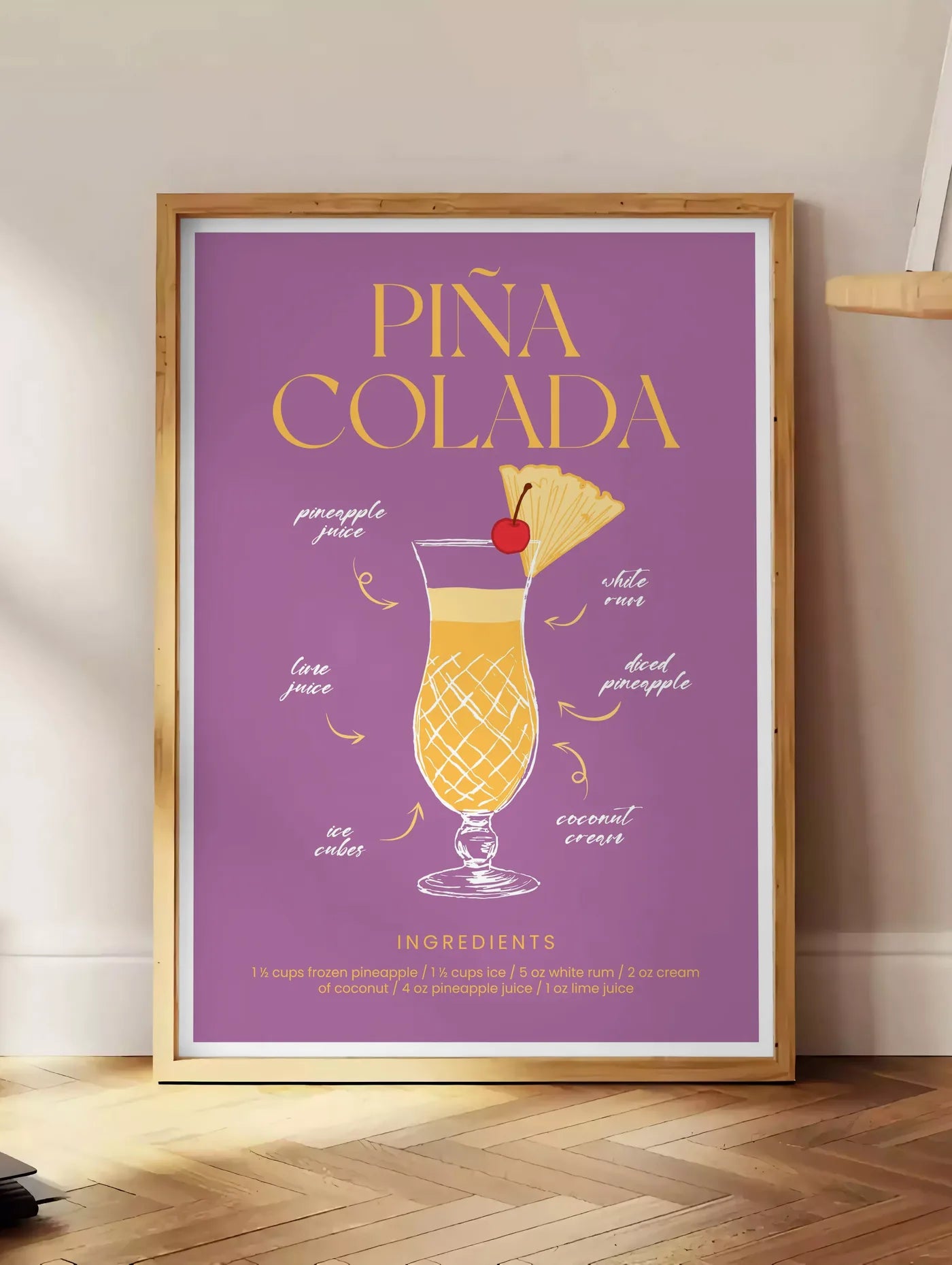 Estampe affiche cocktail Pina Colada, posee au sol dans un coin salon, sur un parquet a chevrons, pres d'une table d'appoint en chene chargee de livres