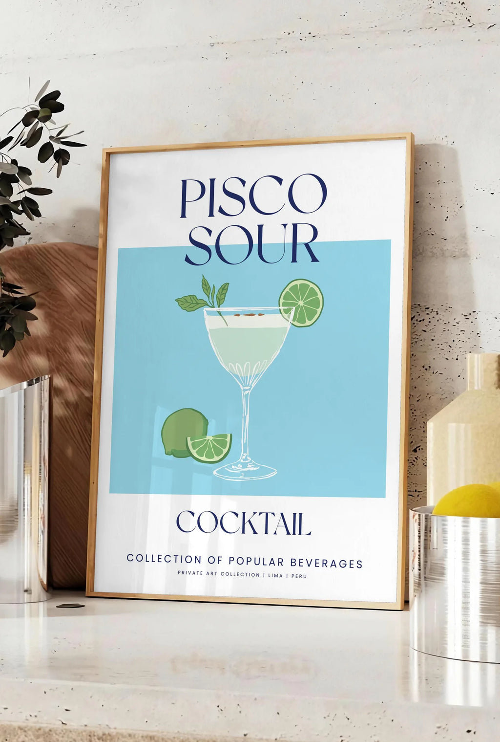 Affiche affiche cocktail Pisco Sour, posee sur un plan de travail de cuisine en pierre claire, entre une branche d'eucalyptus en vase et un bocal de citrons