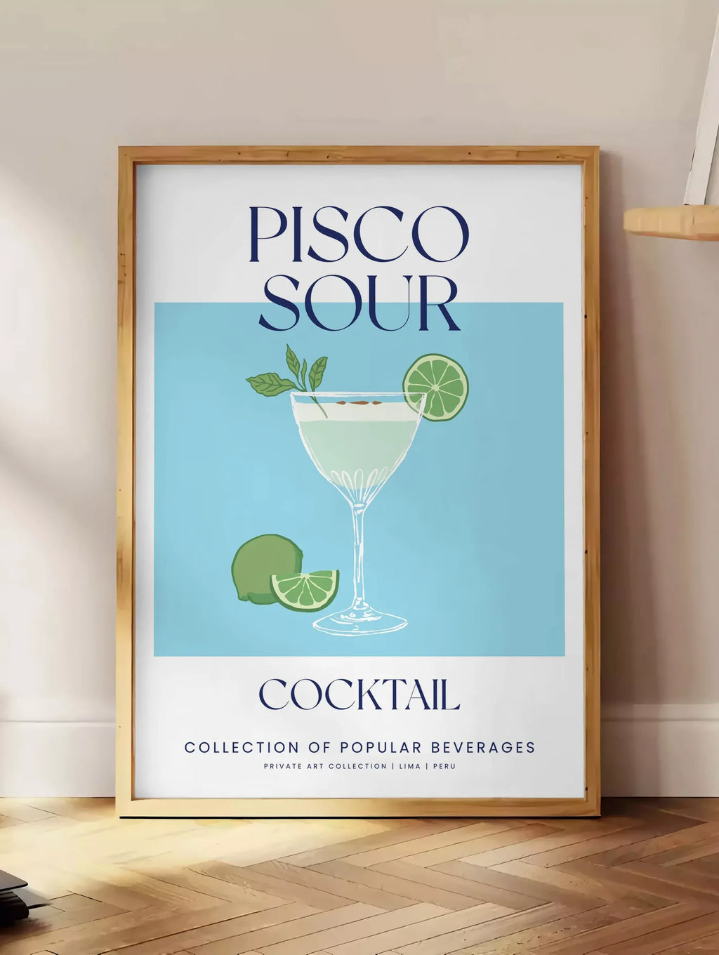 Estampe affiche cocktail Pisco Sour, posee au sol dans un coin salon, sur un parquet a chevrons, pres d'une table d'appoint en chene chargee de livres