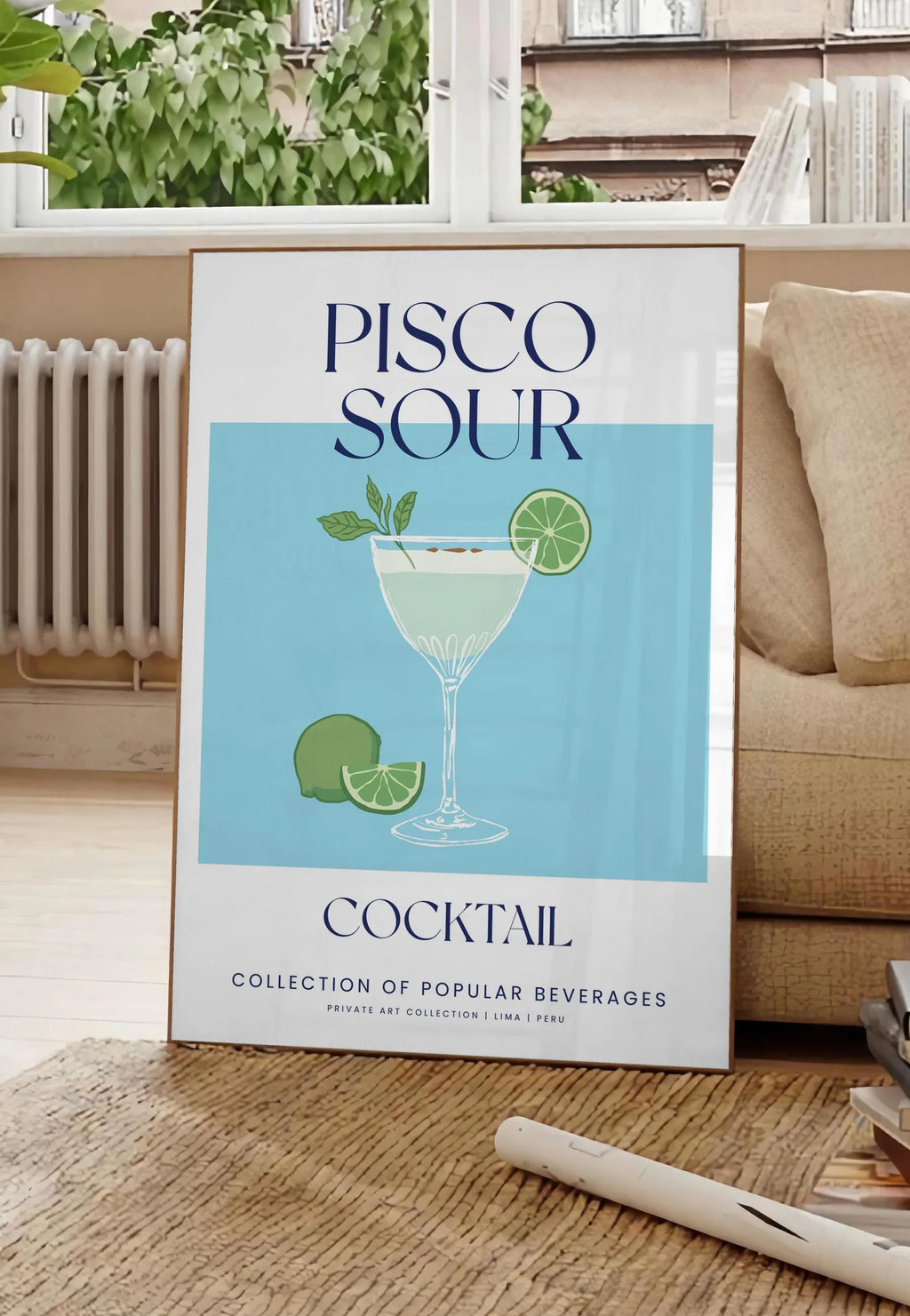 Poster affiche cocktail Pisco Sour, posee au sol dans un salon parisien, devant une grande fenetre donnant sur un immeuble haussmannien, entre un ficus et un canape creme