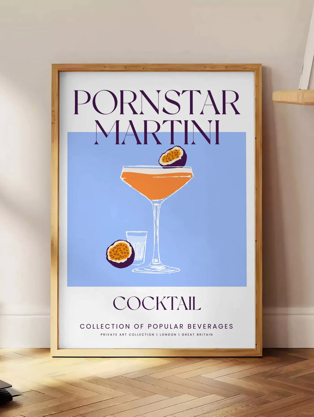 Estampe affiche cocktail Pornstar Martini, posee au sol dans un coin salon, sur un parquet a chevrons, pres d'une table d'appoint en chene chargee de livres