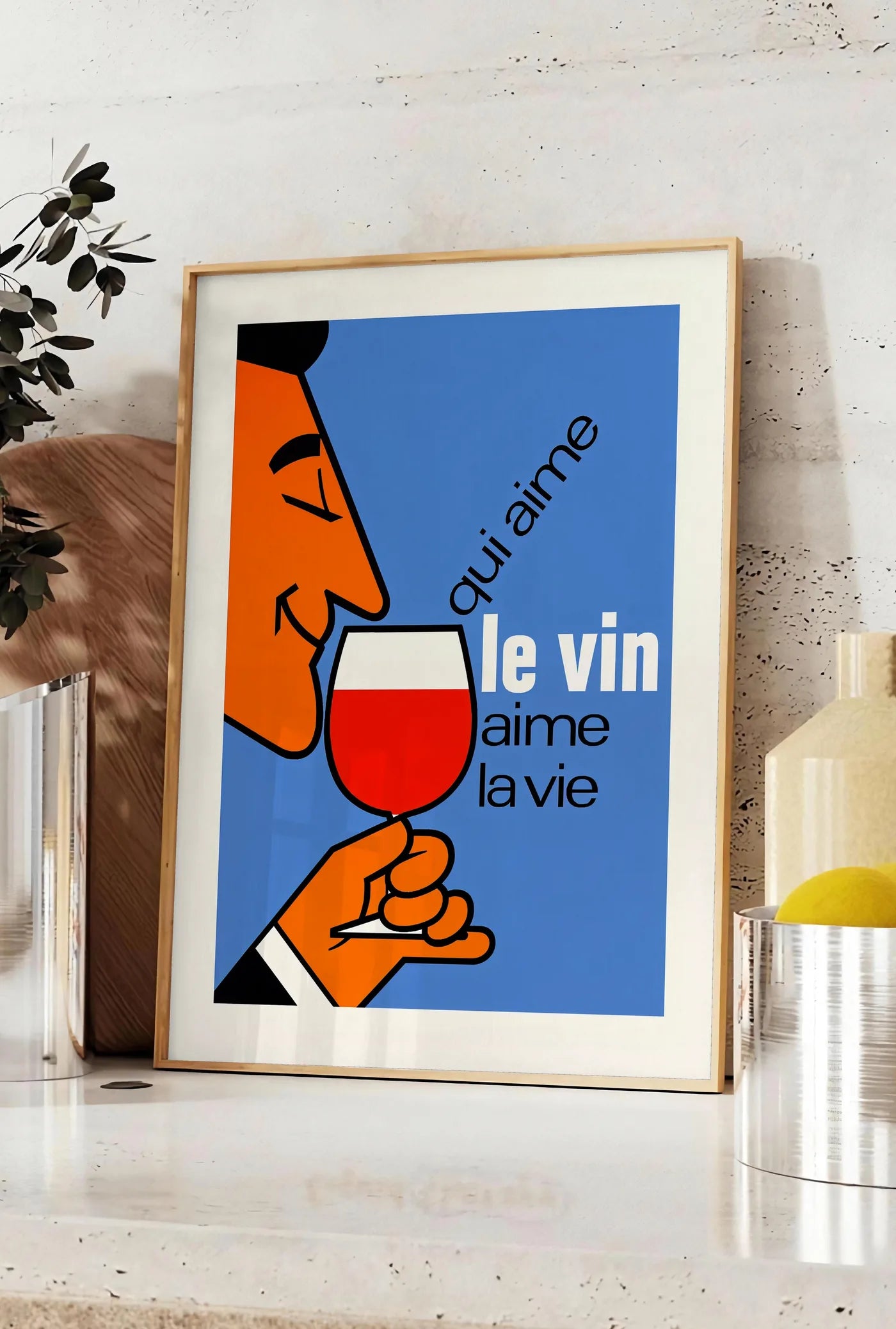 Affiche affiche Qui Aime Le Vin, posee sur un plan de travail de cuisine en pierre claire, entre une branche d'eucalyptus en vase et un bocal de citrons