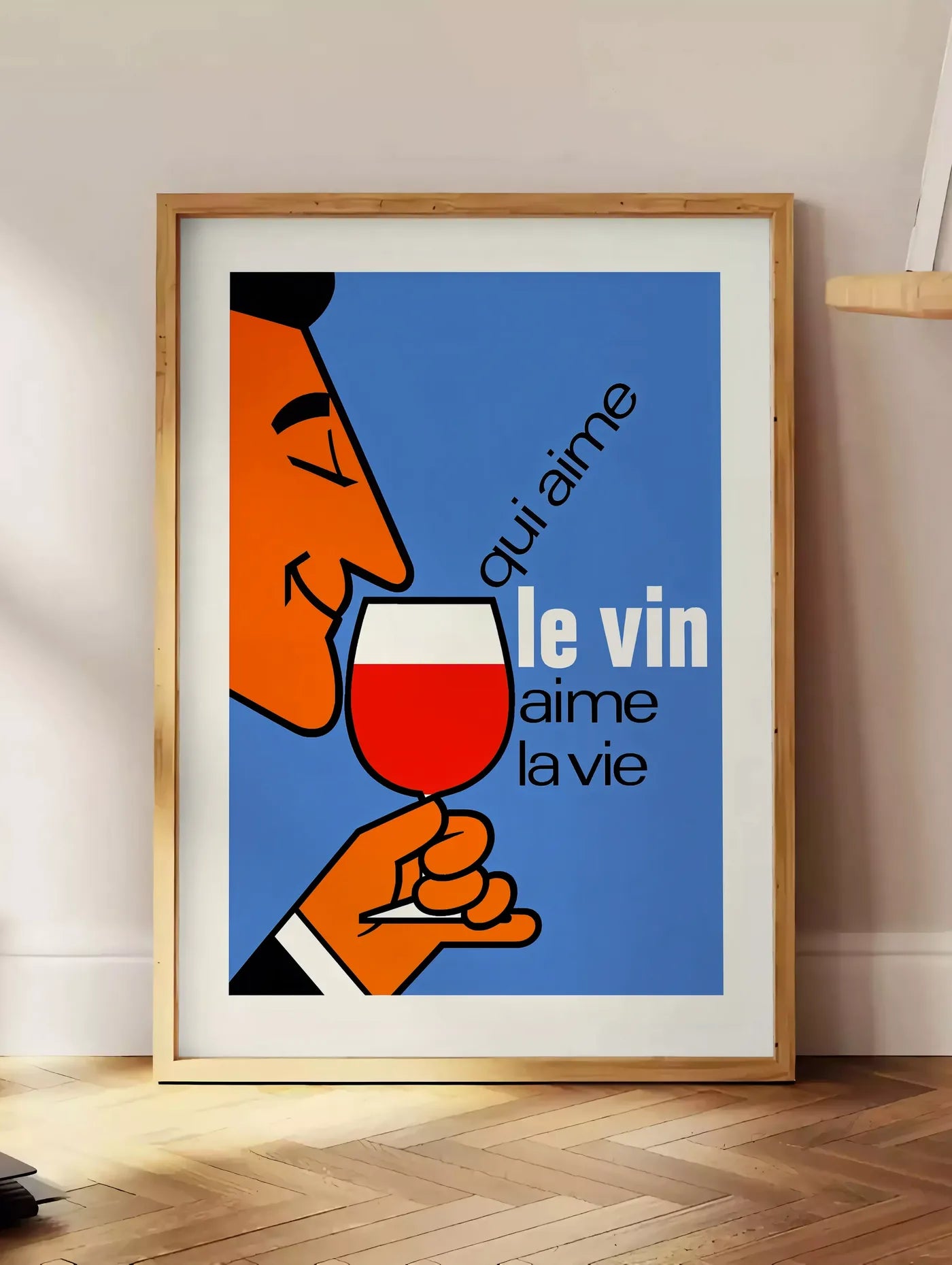Estampe affiche Qui Aime Le Vin, posee au sol dans un coin salon, sur un parquet a chevrons, pres d'une table d'appoint en chene chargee de livres