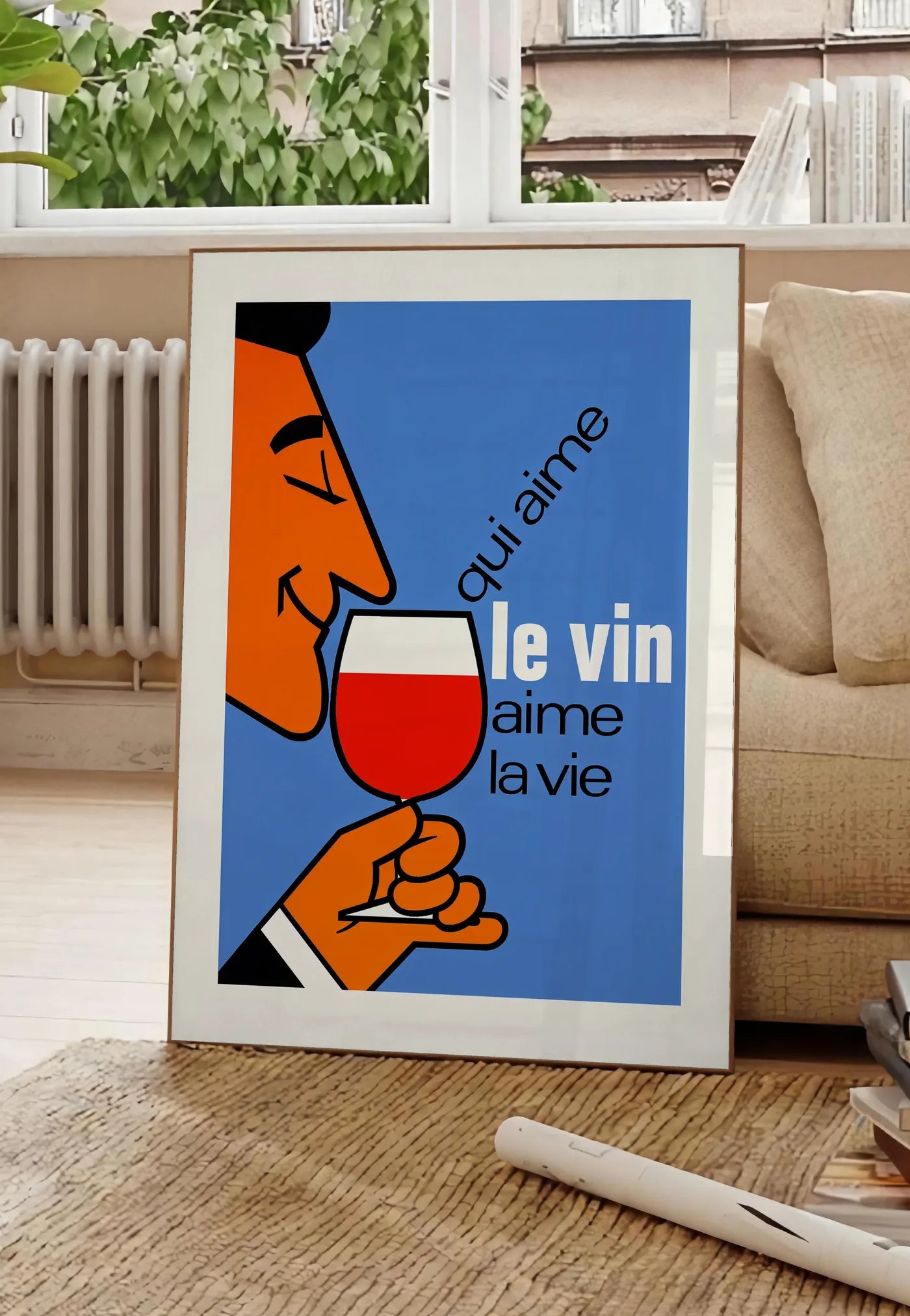 Poster affiche Qui Aime Le Vin, posee au sol dans un salon parisien, devant une grande fenetre donnant sur un immeuble haussmannien, entre un ficus et un canape creme
