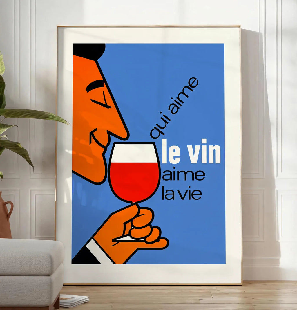 Reproduction affiche Qui Aime Le Vin, posee au sol dans un salon minimaliste clair, a cote d'un canape beige et d'une plante verte, sur un parquet clair
