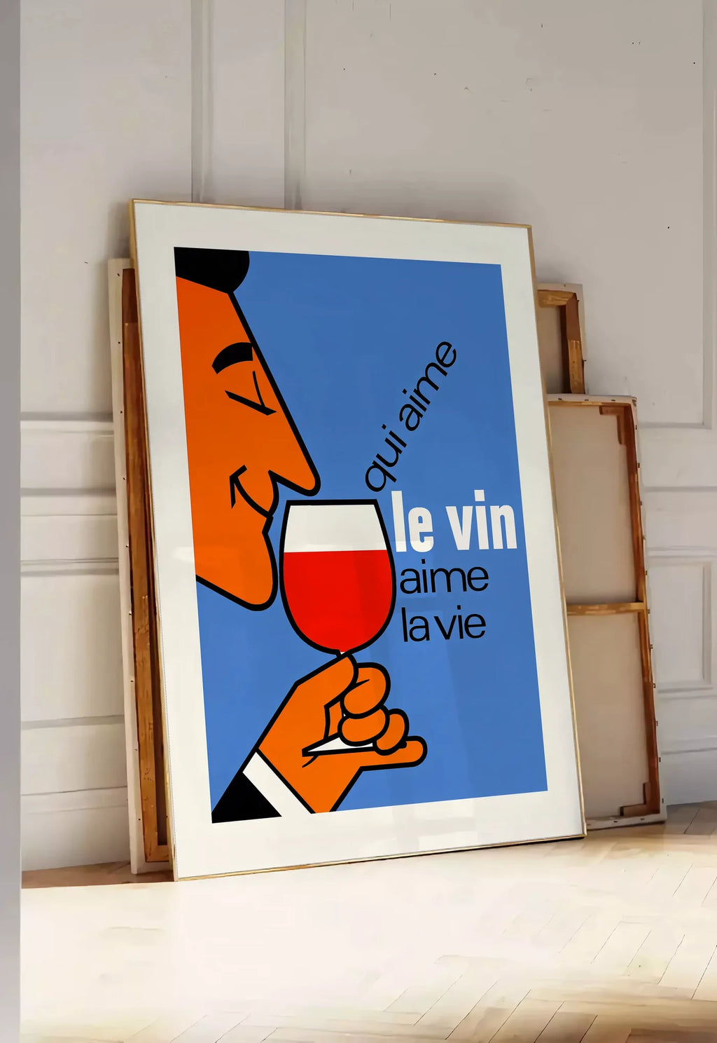 Tirage encadrable affiche Qui Aime Le Vin, posee au sol dans une entree lumineuse, contre un mur a panneaux blancs, a cote de chassis en bois empiles