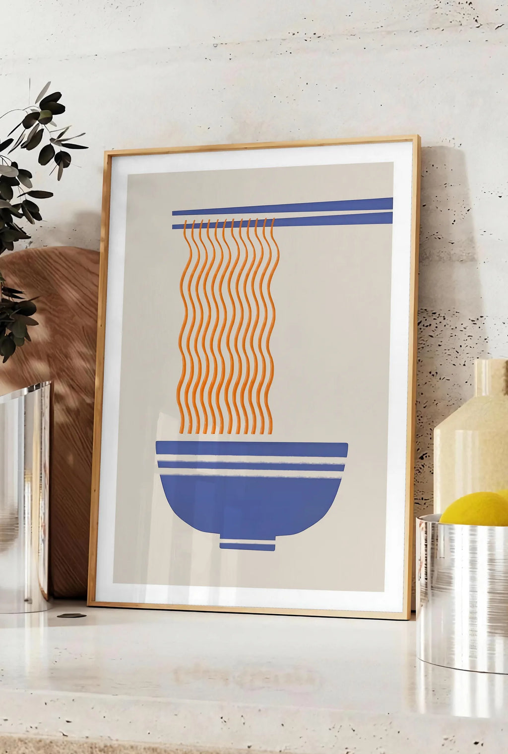 Affiche affiche cuisine ramen, posee sur un plan de travail de cuisine en pierre claire, entre une branche d'eucalyptus en vase et un bocal de citrons