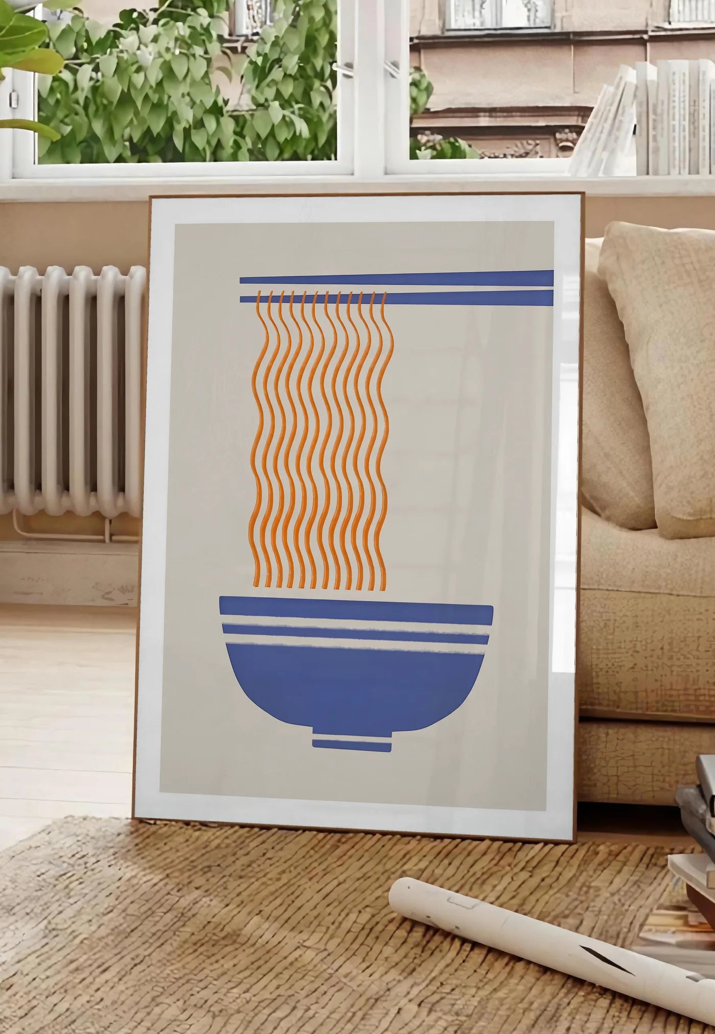 Poster affiche cuisine ramen, posee au sol dans un salon parisien, devant une grande fenetre donnant sur un immeuble haussmannien, entre un ficus et un canape creme