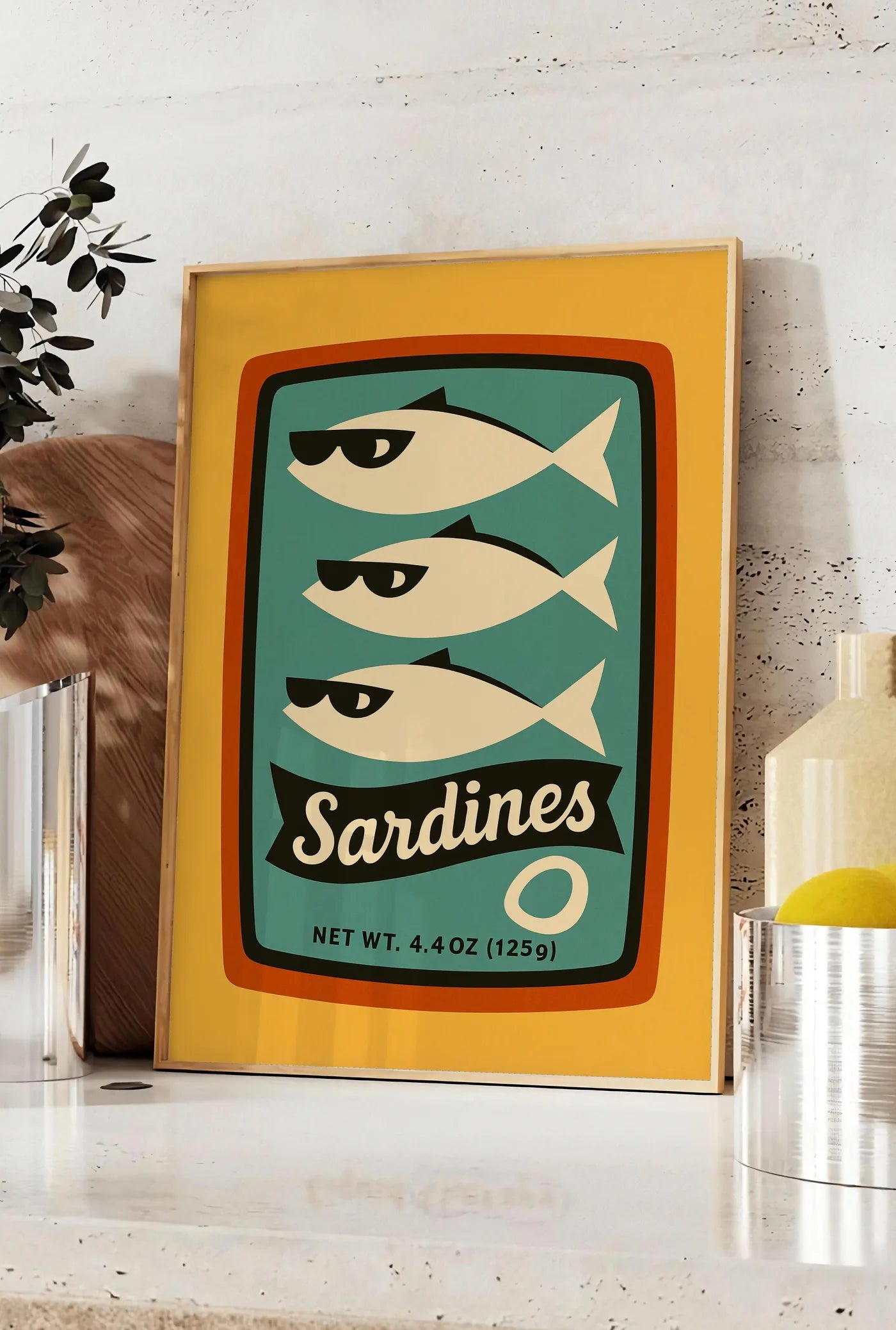 Affiche affiche cuisine sardines, posee sur un plan de travail de cuisine en pierre claire, entre une branche d'eucalyptus en vase et un bocal de citrons