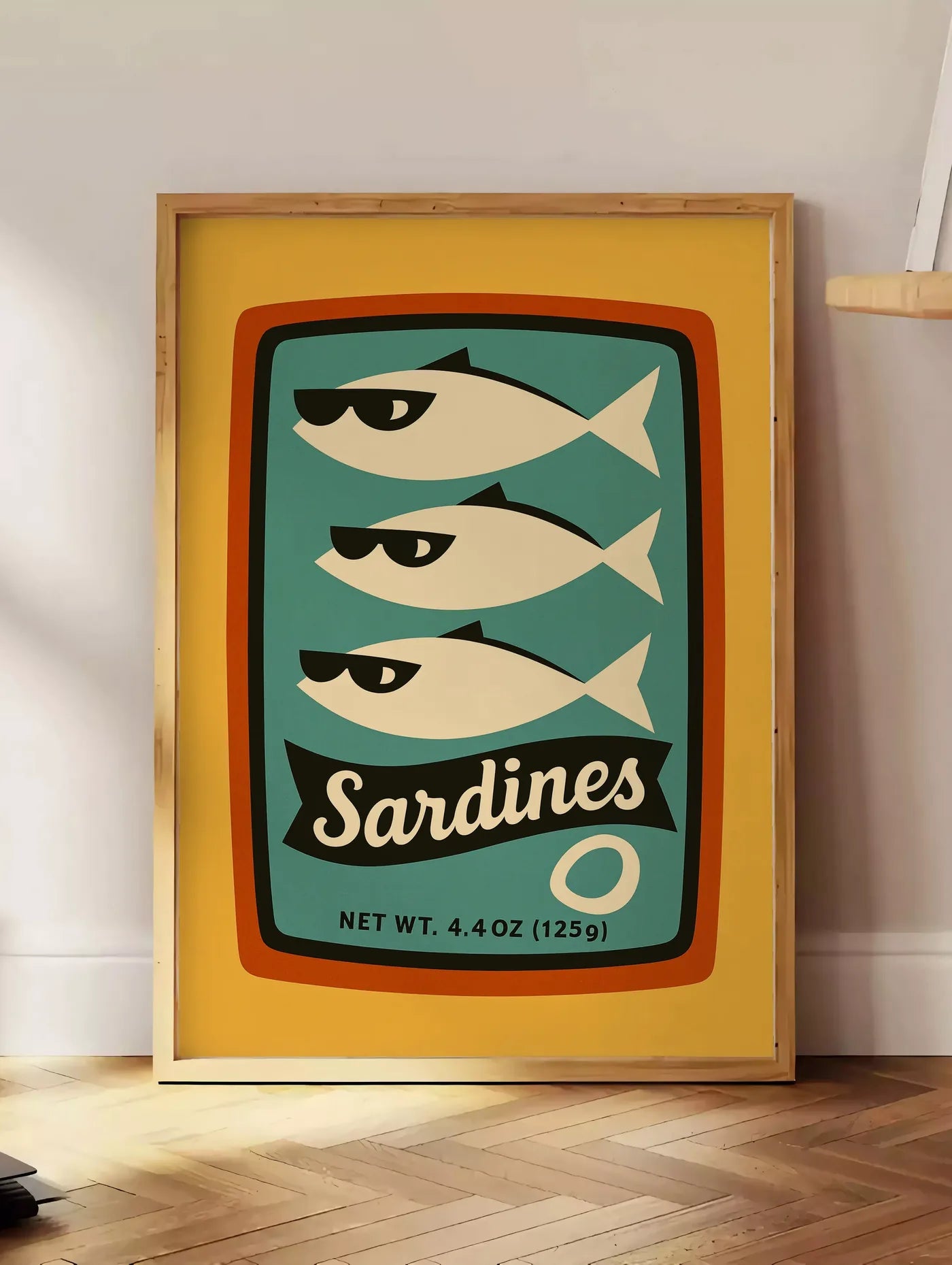 Estampe affiche cuisine sardines, posee au sol dans un coin salon, sur un parquet a chevrons, pres d'une table d'appoint en chene chargee de livres