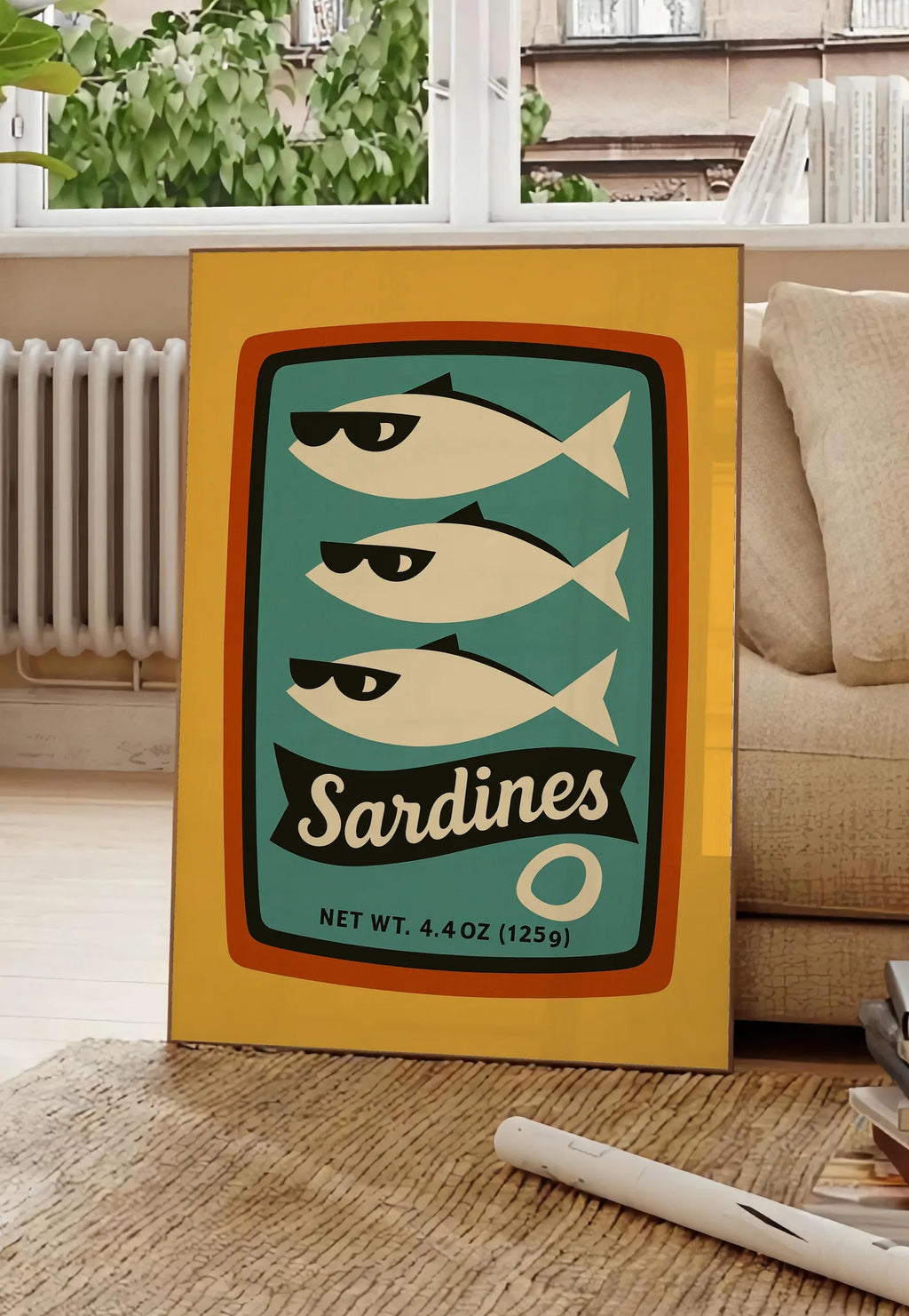 Poster affiche cuisine sardines, posee au sol dans un salon parisien, devant une grande fenetre donnant sur un immeuble haussmannien, entre un ficus et un canape creme
