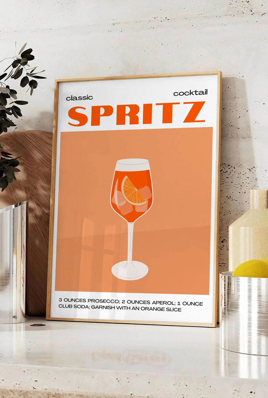 Affiche affiche cocktail Spritz, posee sur un plan de travail de cuisine en pierre claire, entre une branche d'eucalyptus en vase et un bocal de citrons