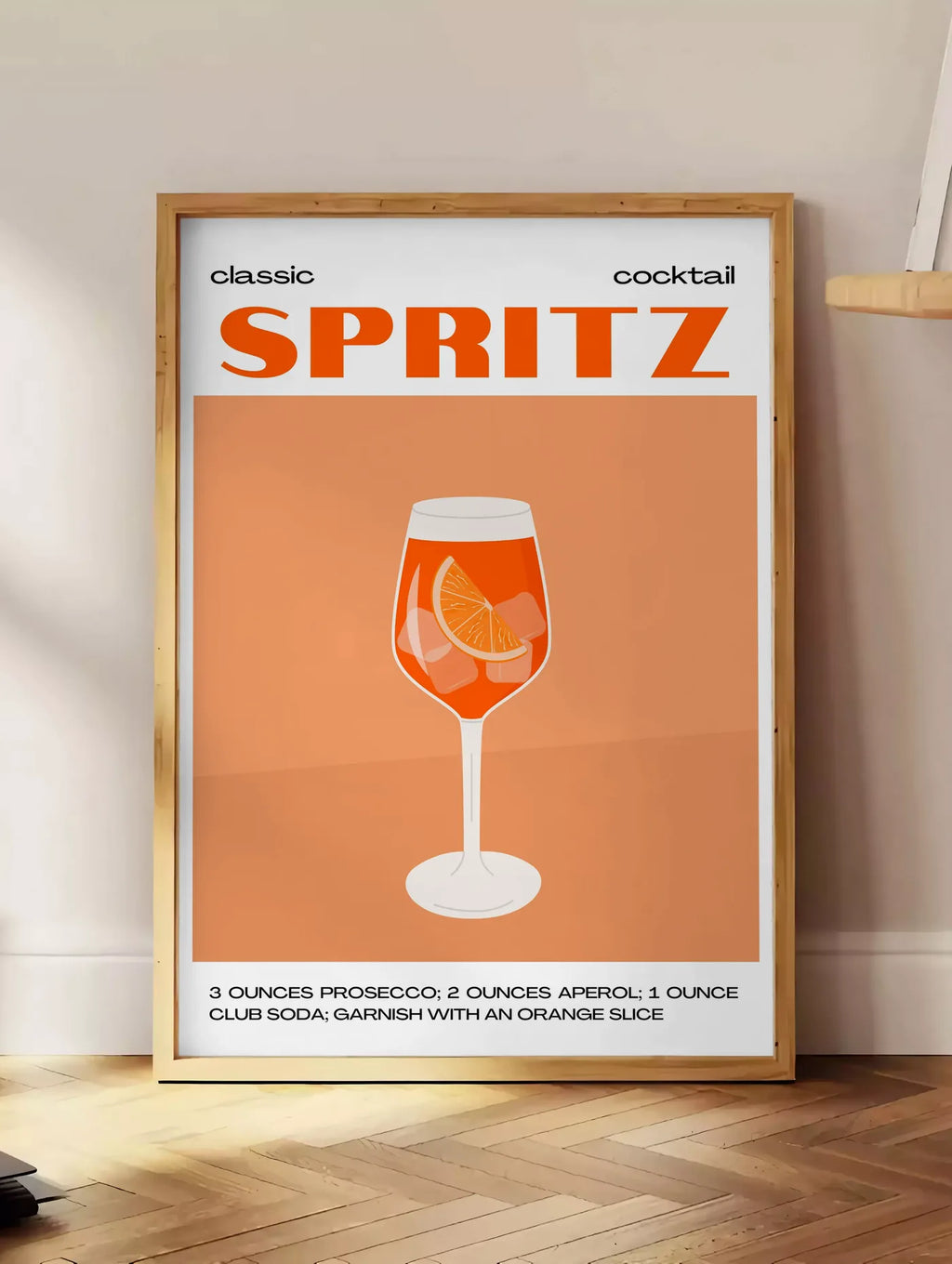 Estampe affiche cocktail Spritz, posee au sol dans un coin salon, sur un parquet a chevrons, pres d'une table d'appoint en chene chargee de livres