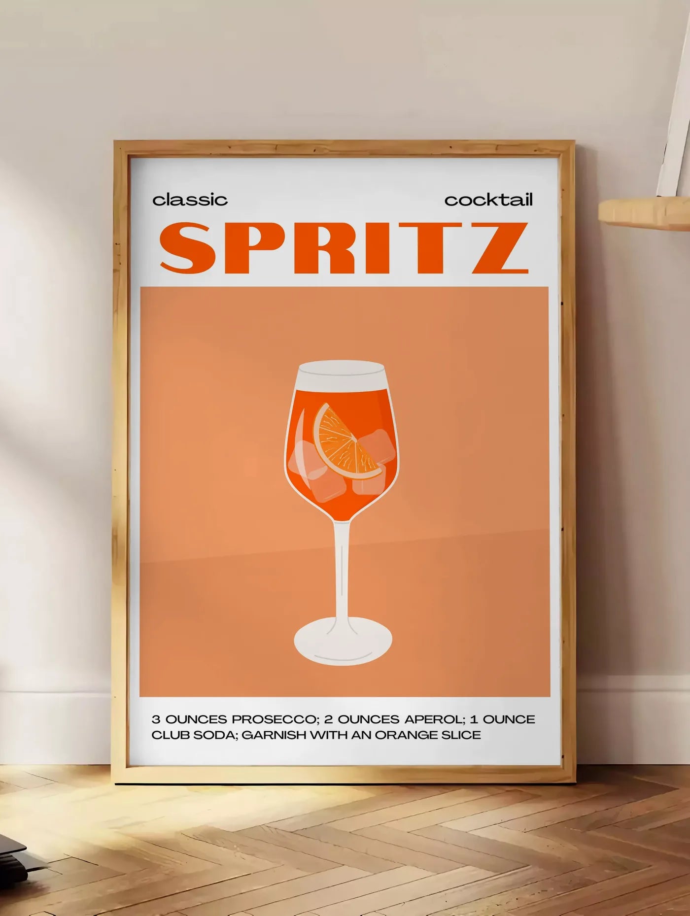 Estampe affiche cocktail Spritz, posee au sol dans un coin salon, sur un parquet a chevrons, pres d'une table d'appoint en chene chargee de livres