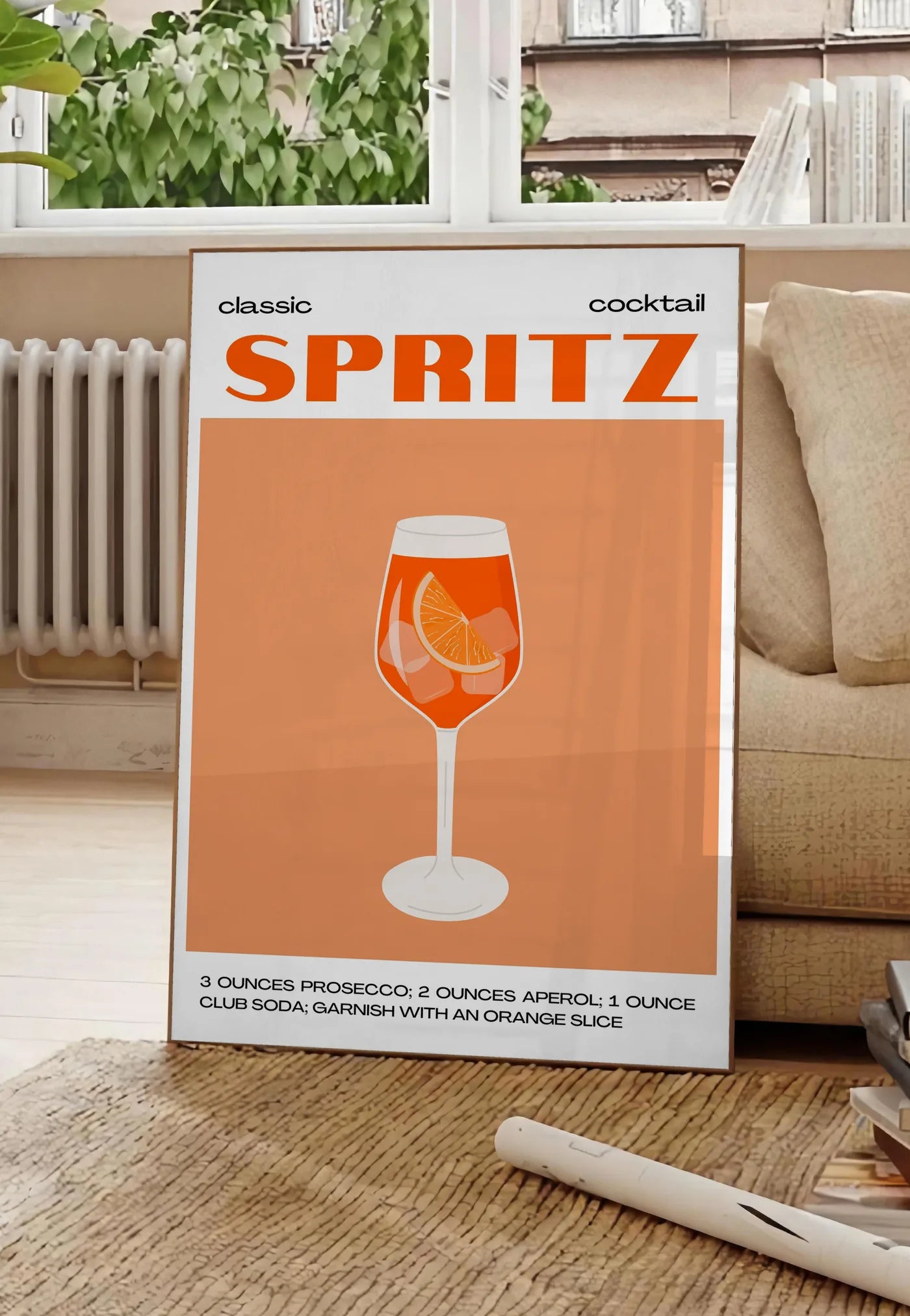 Poster affiche cocktail Spritz, posee au sol dans un salon parisien, devant une grande fenetre donnant sur un immeuble haussmannien, entre un ficus et un canape creme