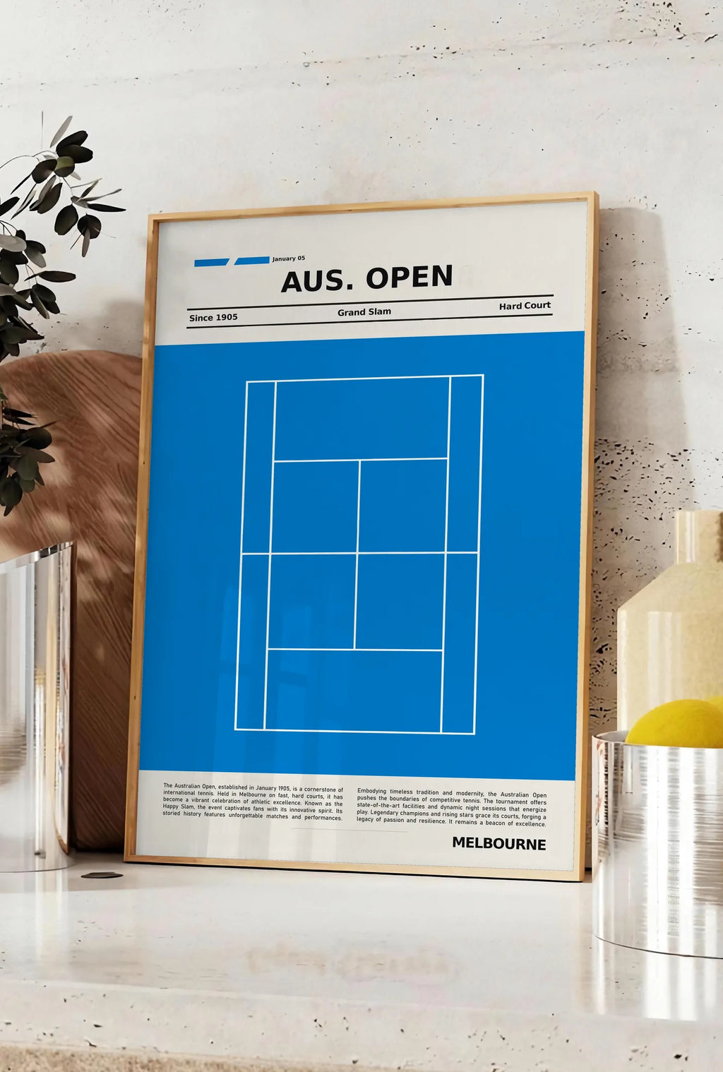 Affiche court de tennis stylisé, posee sur un plan de travail de cuisine en pierre claire, entre une branche d'eucalyptus en vase et un bocal de citrons