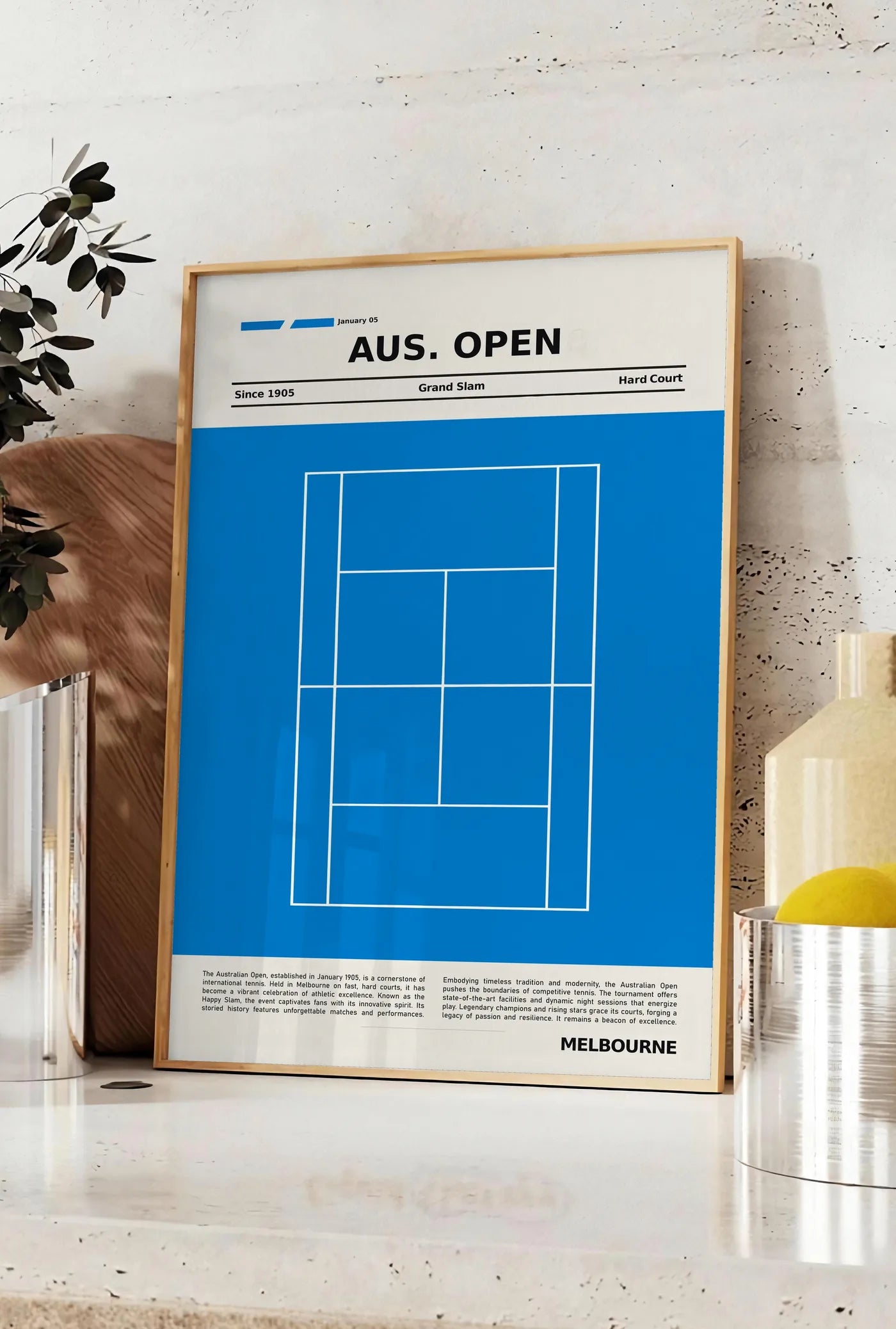Affiche court de tennis stylisé, posee sur un plan de travail de cuisine en pierre claire, entre une branche d'eucalyptus en vase et un bocal de citrons