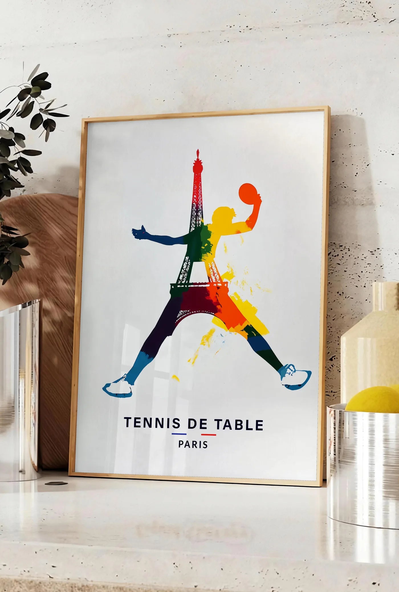 Affiche joueur de tennis de table bondissant, posee sur un plan de travail de cuisine en pierre claire, entre une branche d'eucalyptus en vase et un bocal de citrons
