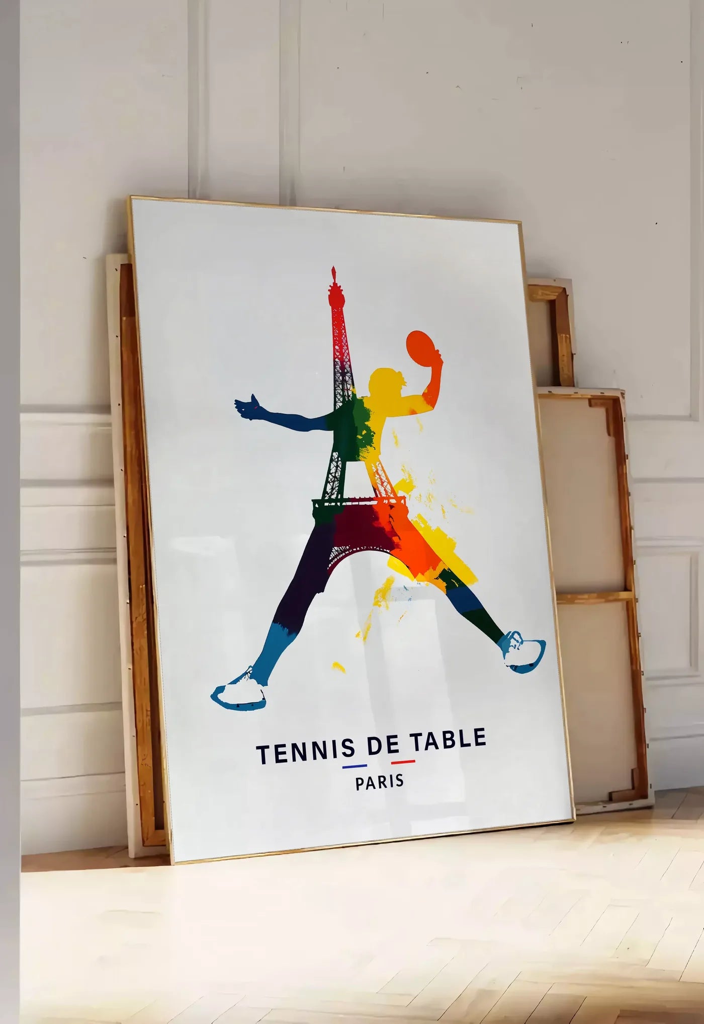 Tirage encadrable joueur de tennis de table bondissant, posee au sol dans une entree lumineuse, contre un mur a panneaux blancs, a cote de chassis en bois empiles