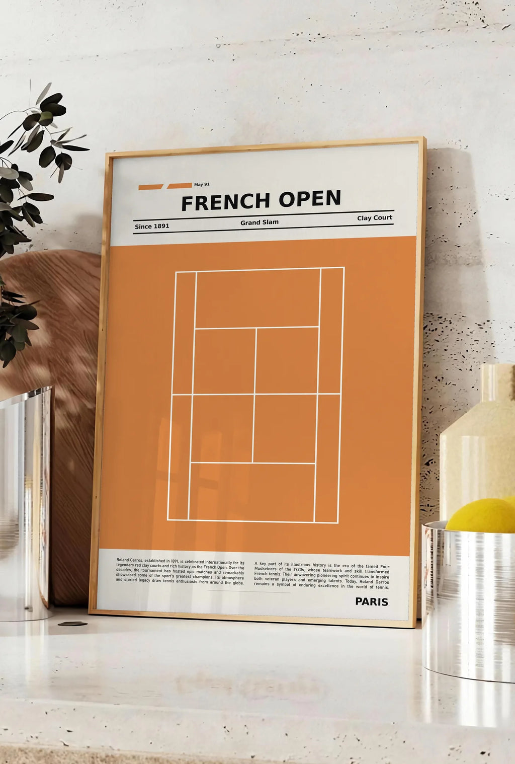 Affiche court de tennis stylisé en terre battue avec titre FRENCH OPEN, posee sur un plan de travail de cuisine en pierre claire, entre une branche d'eucalyptus en vase et un bocal de citrons