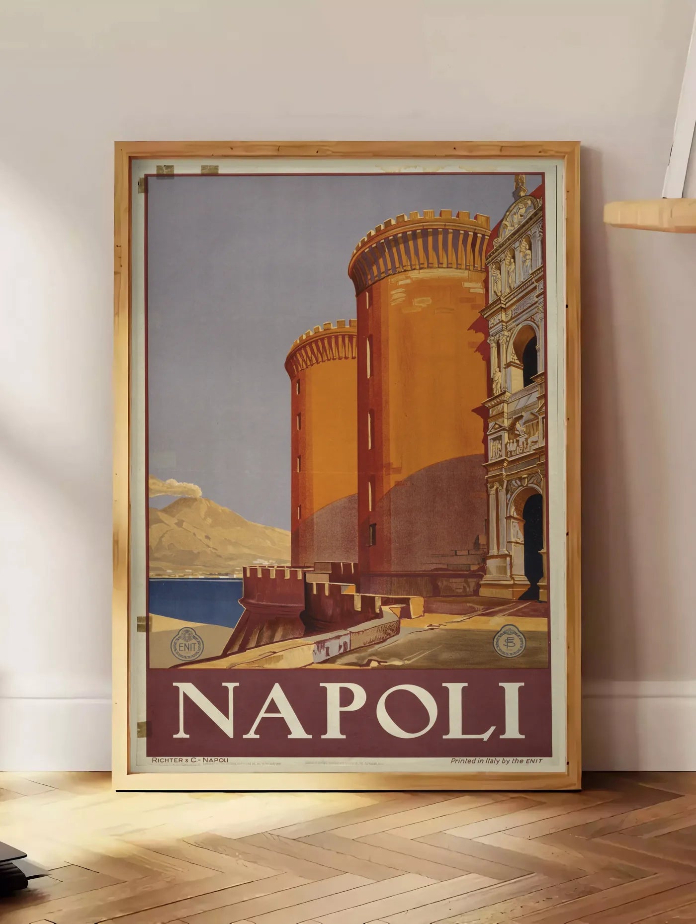 Estampe affiche Napoli vintage ENIT, posee au sol dans un coin salon, sur un parquet a chevrons, pres d'une table d'appoint en chene chargee de livres
