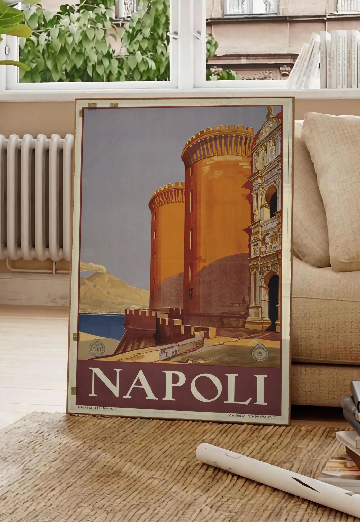 Poster affiche Napoli vintage ENIT, posee au sol dans un salon parisien, devant une grande fenetre donnant sur un immeuble haussmannien, entre un ficus et un canape creme