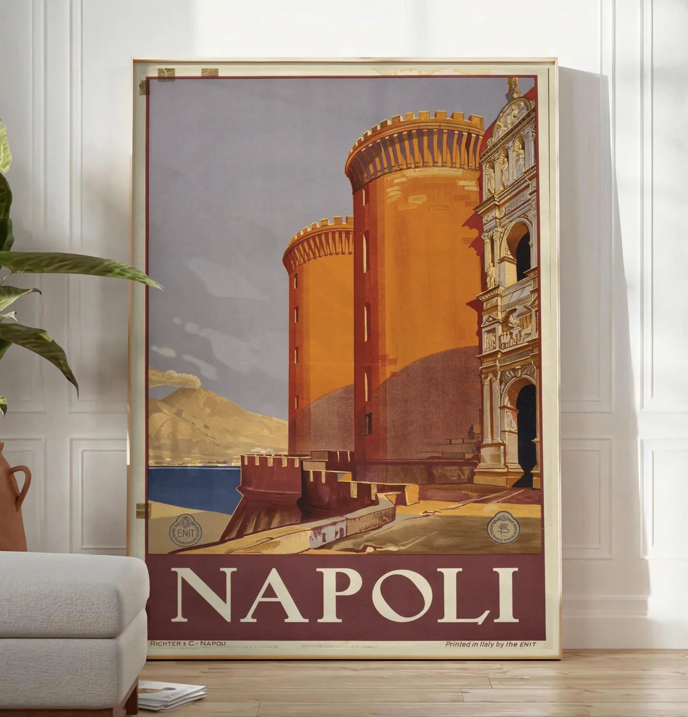 Reproduction affiche Napoli vintage ENIT, posee au sol dans un salon minimaliste clair, a cote d'un canape beige et d'une plante verte, sur un parquet clair