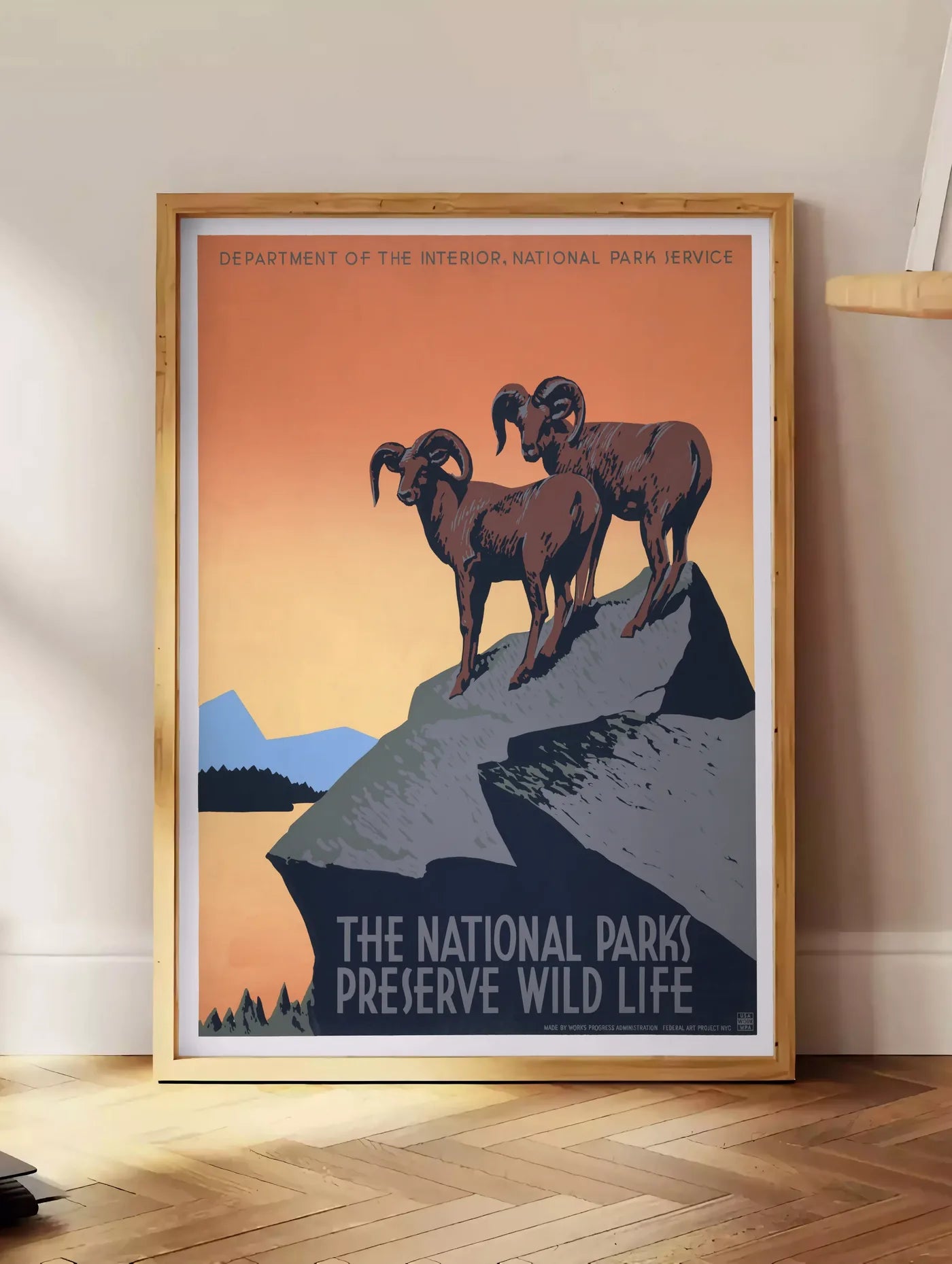 Estampe affiche mouflons National Parks vintage, posee au sol dans un coin salon, sur un parquet a chevrons, pres d'une table d'appoint en chene chargee de livres